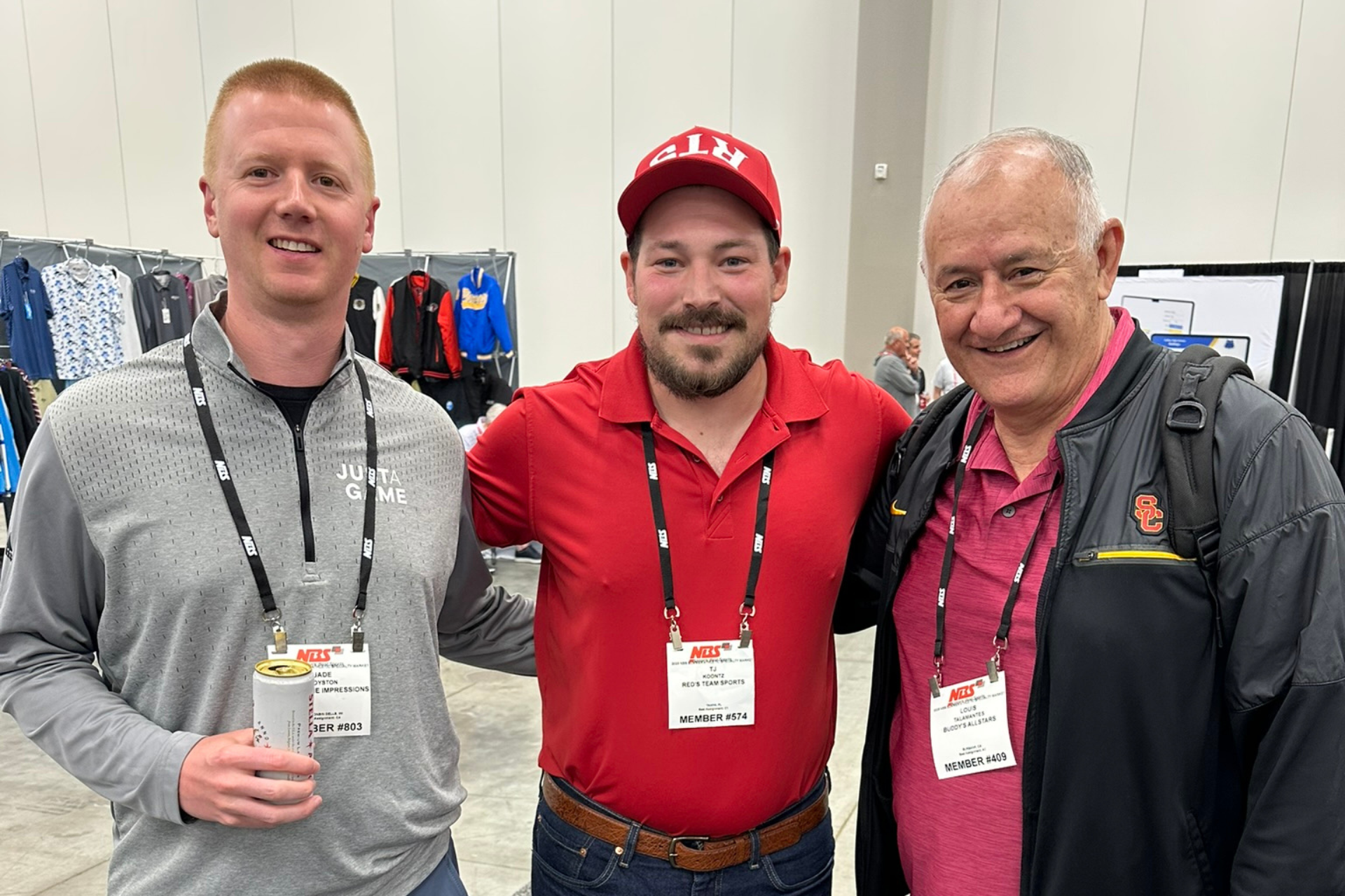 Three men smiling at an indoor event, wearing lanyards and casual clothes, with clothing racks behind them