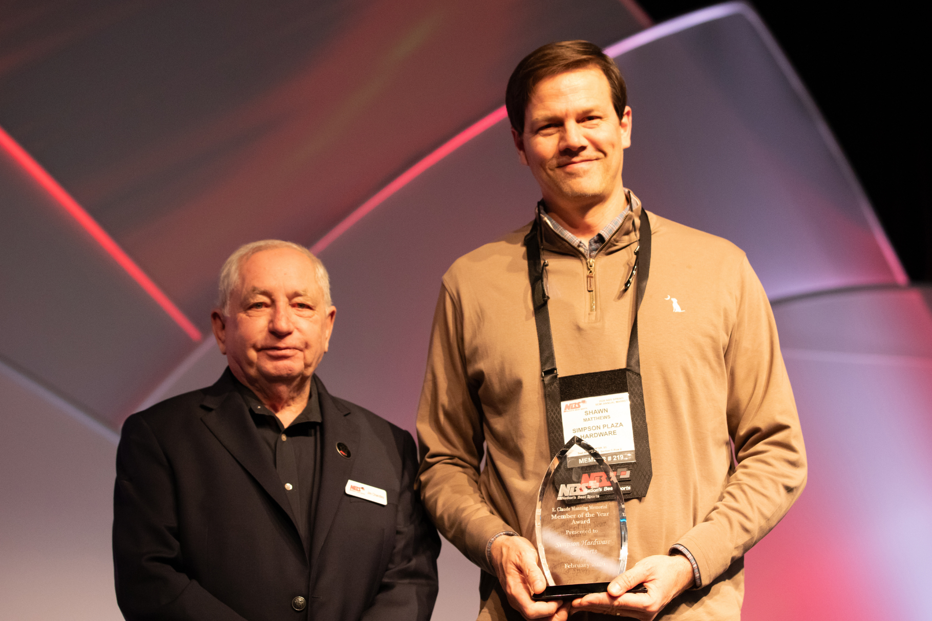 Two men on stage holding an award trophy in front of a lit backdrop