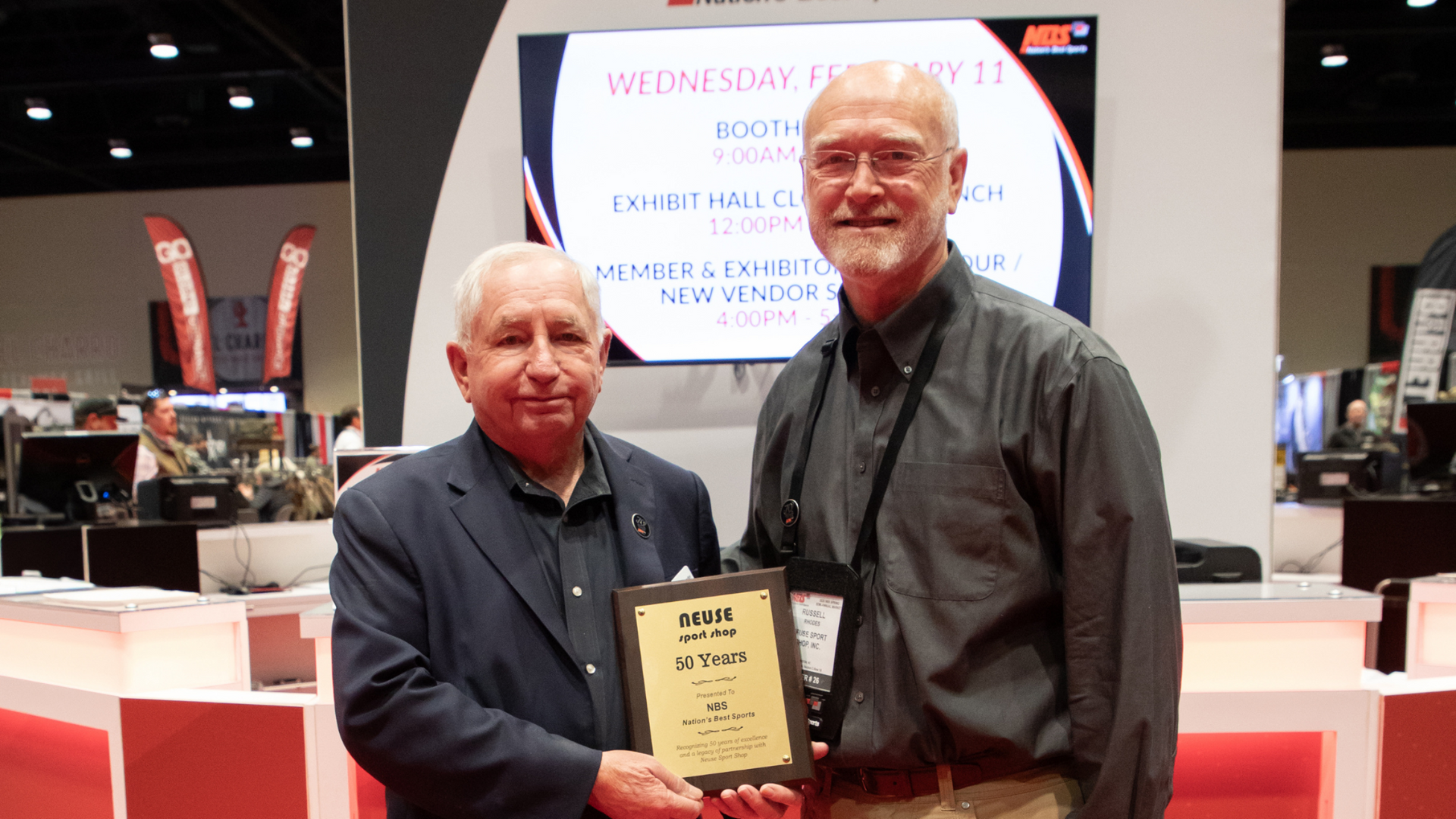 Two men holding a plaque at a trade show booth, with a presentation screen in the background.
