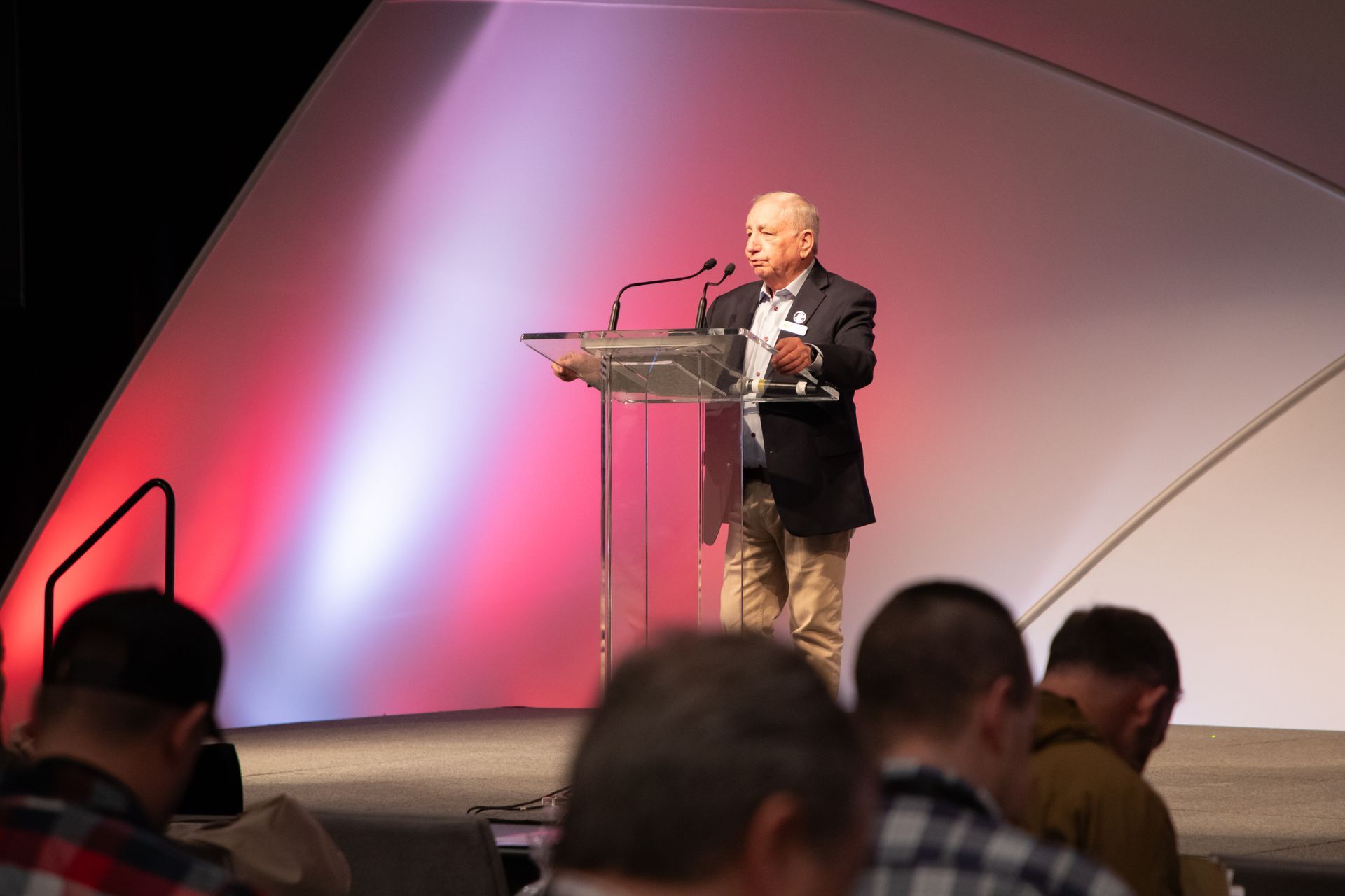 Speaker at a podium on a stage with pink and white lighting, seen above an audience.