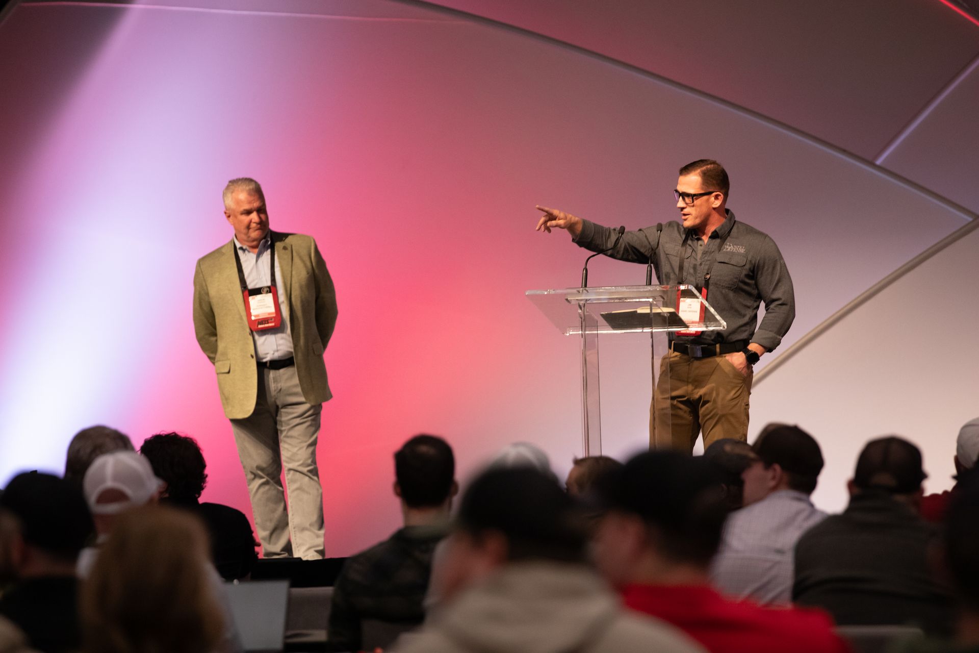 Two speakers on a stage, one pointing at a podium in front of an audience.