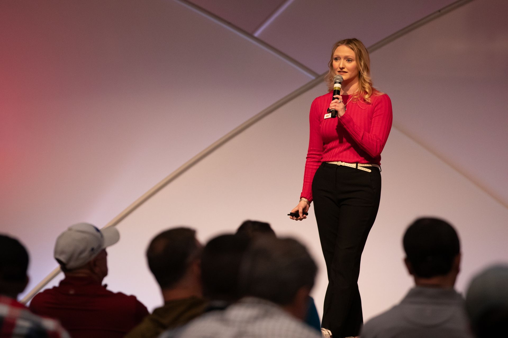Woman in bright red top speaking on stage with a microphone, audience blurred in foreground