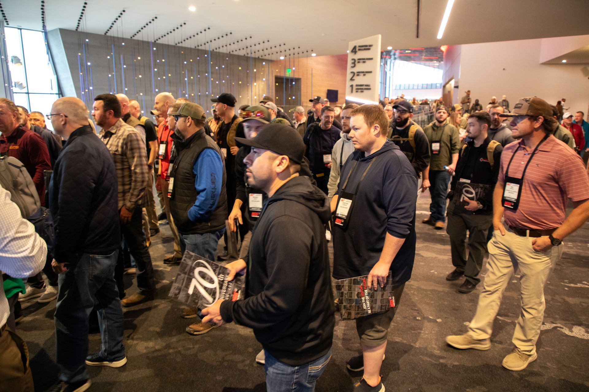 Crowd of people standing in a lobby or conference hall, some holding cameras and wearing lanyards.