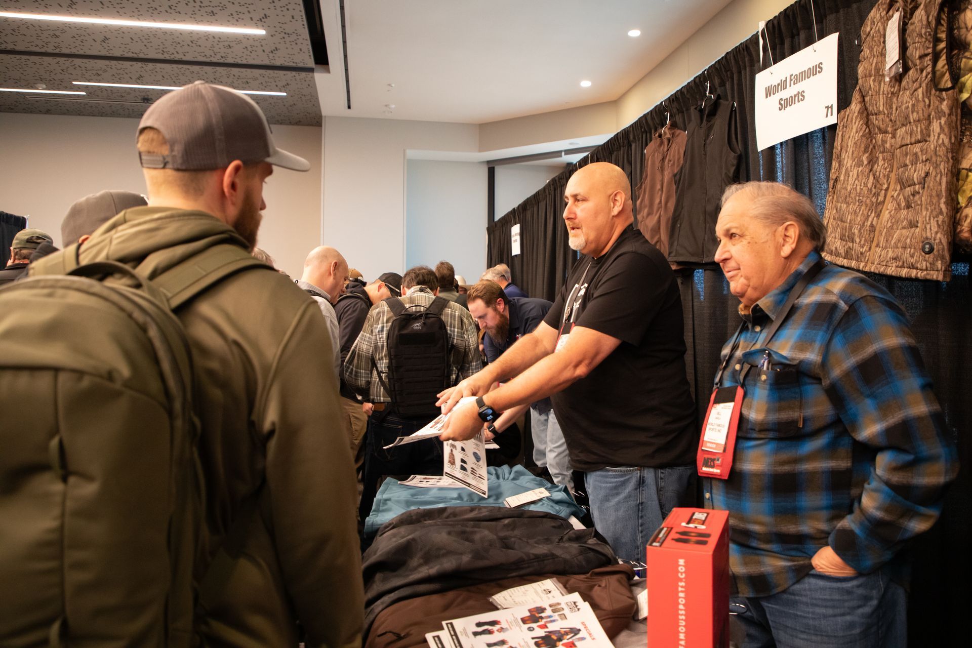 People gathered at a trade show booth, with two men behind a table speaking to attendees.