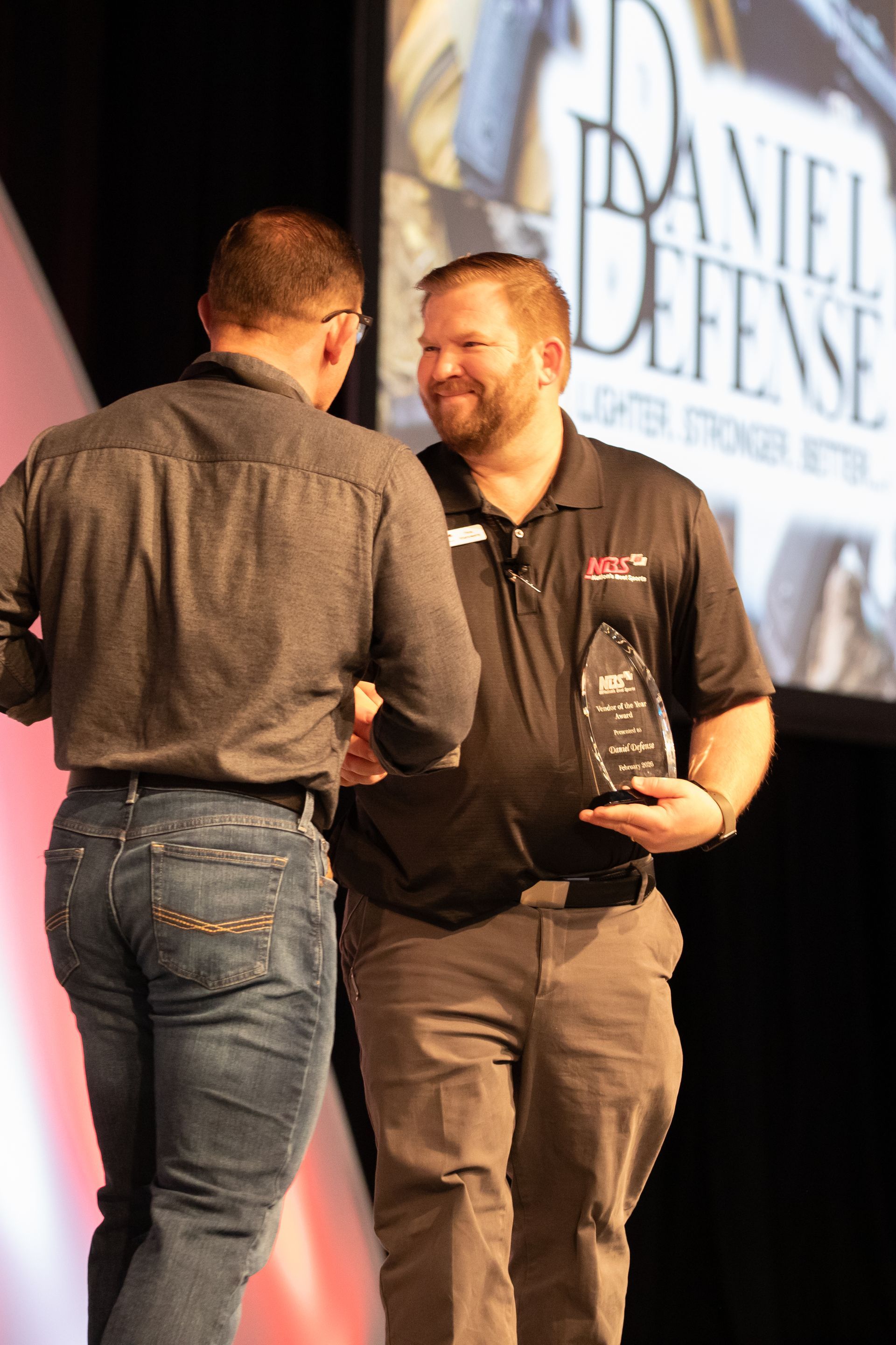 Two men onstage shake hands, one holding a trophy, with a Daniel Defense screen in the background.