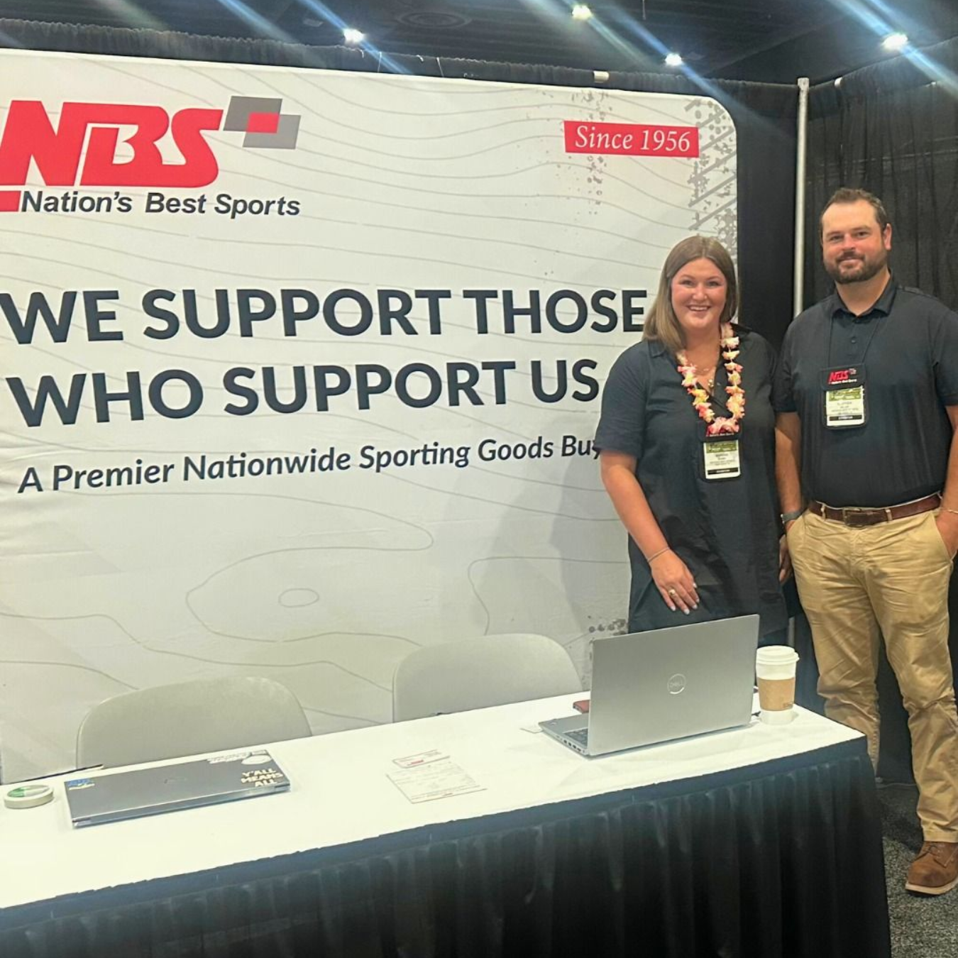 Trade show booth for NBS with two staff standing behind a table and a banner reading “We support those who support us.”