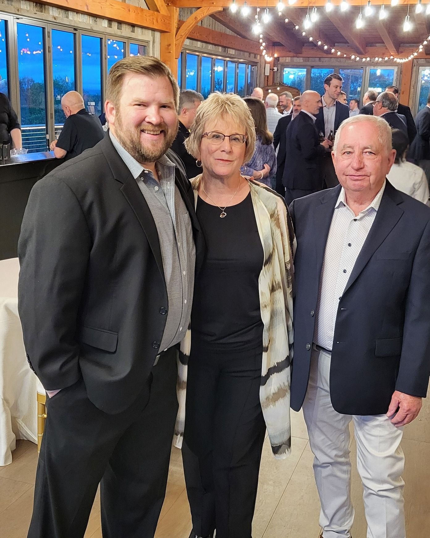 Three people standing together at an indoor event with string lights and guests in the background.