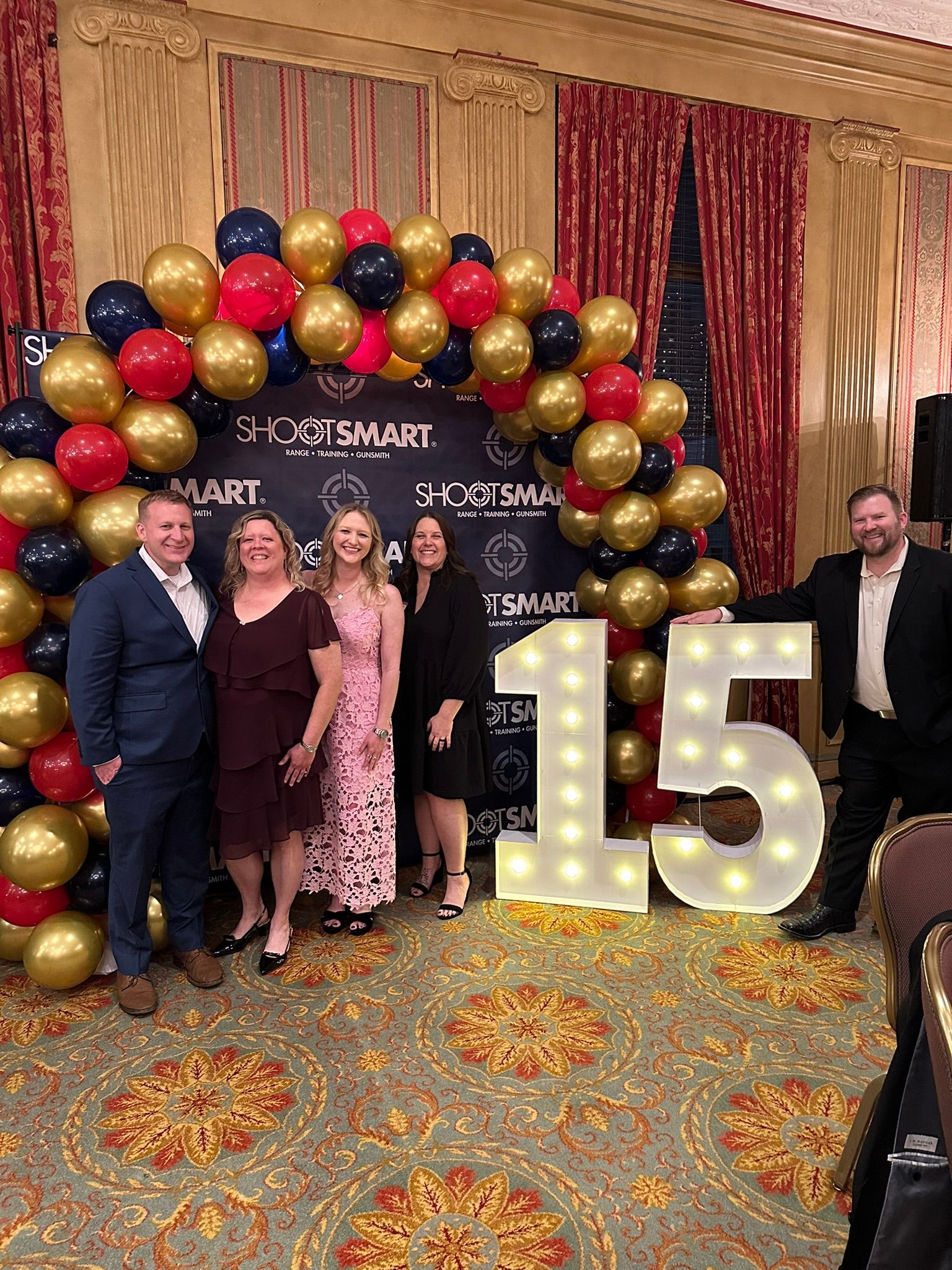 Five people posing at a 15th business anniversary celebration with gold and black balloons and large lit “15” numbers