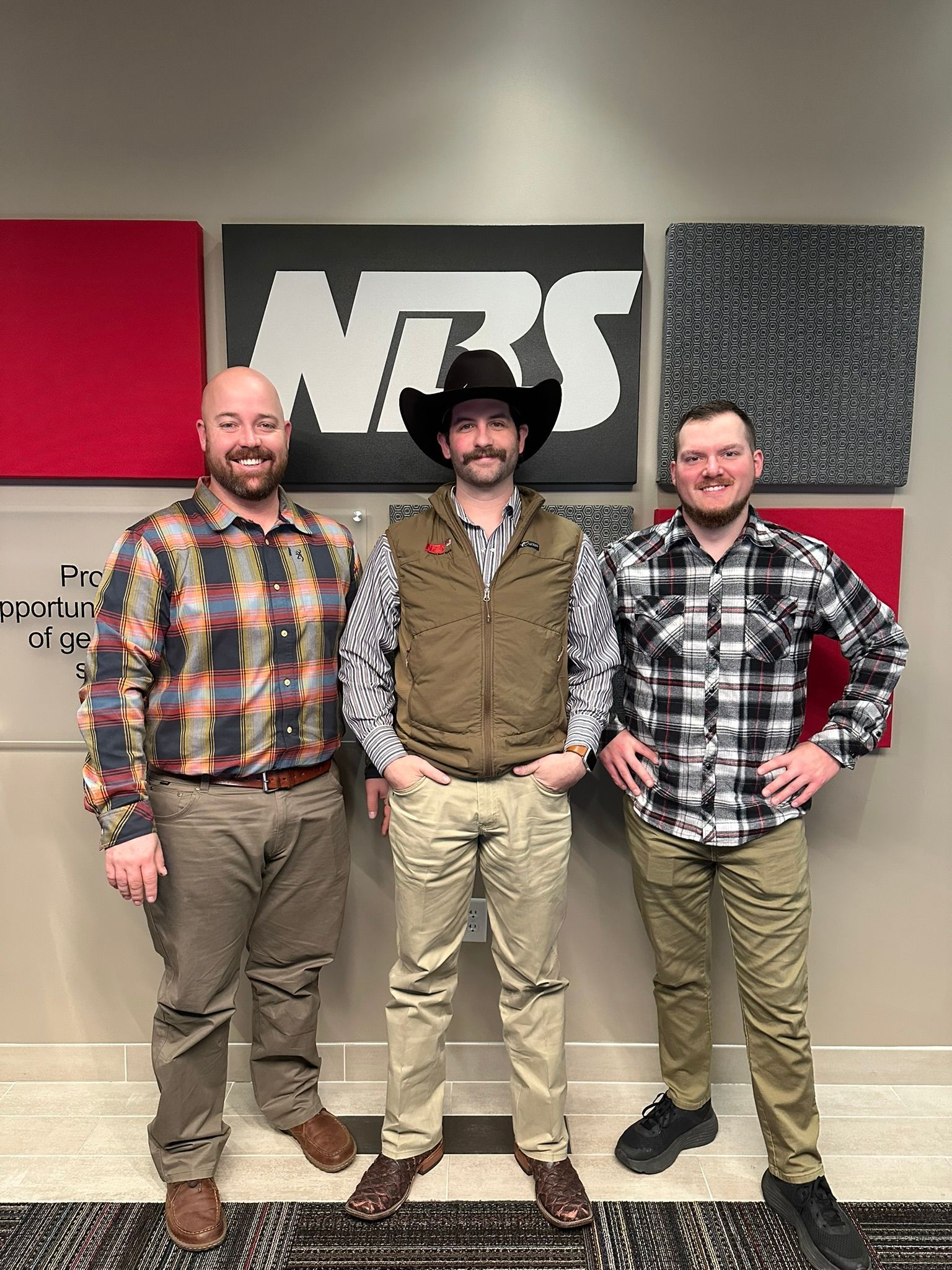 Three men posing in front of a wall with a large NBS logo, wearing plaid shirts and cowboy hats.
