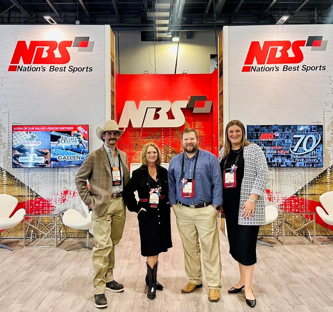 Four people posing at an NBS trade show booth with red-and-white branding.