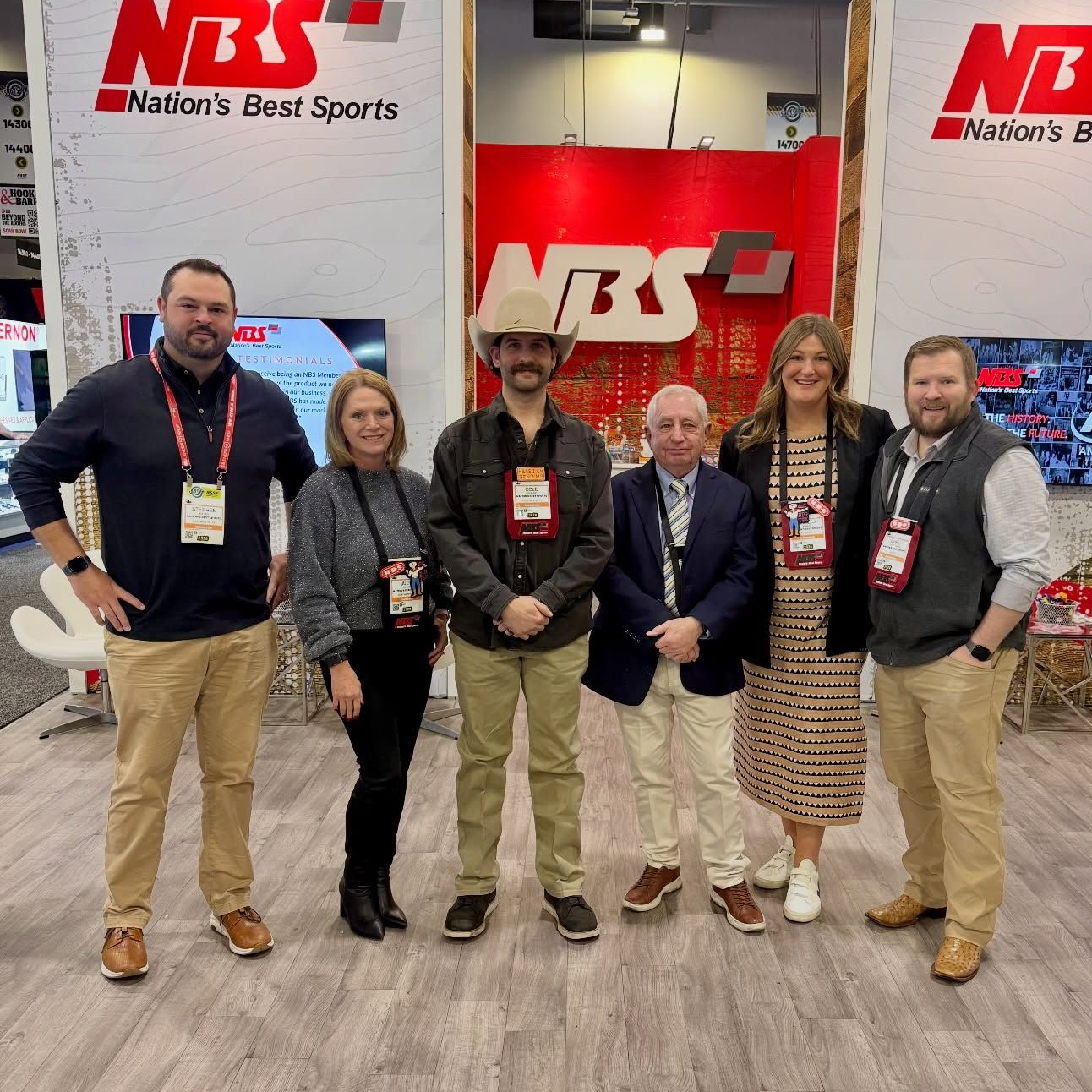 Five people posing at a Nation’s Best Sports booth with NBS banners and red backdrop at a trade show.