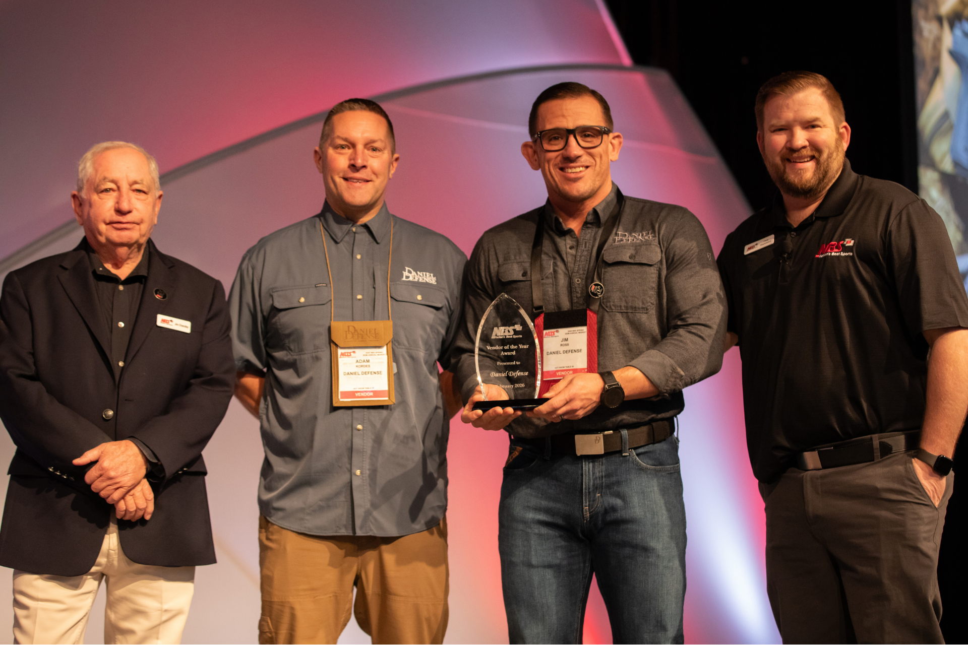 Four men posing on a stage, one holding an award plaque, with pink and black lighting behind them