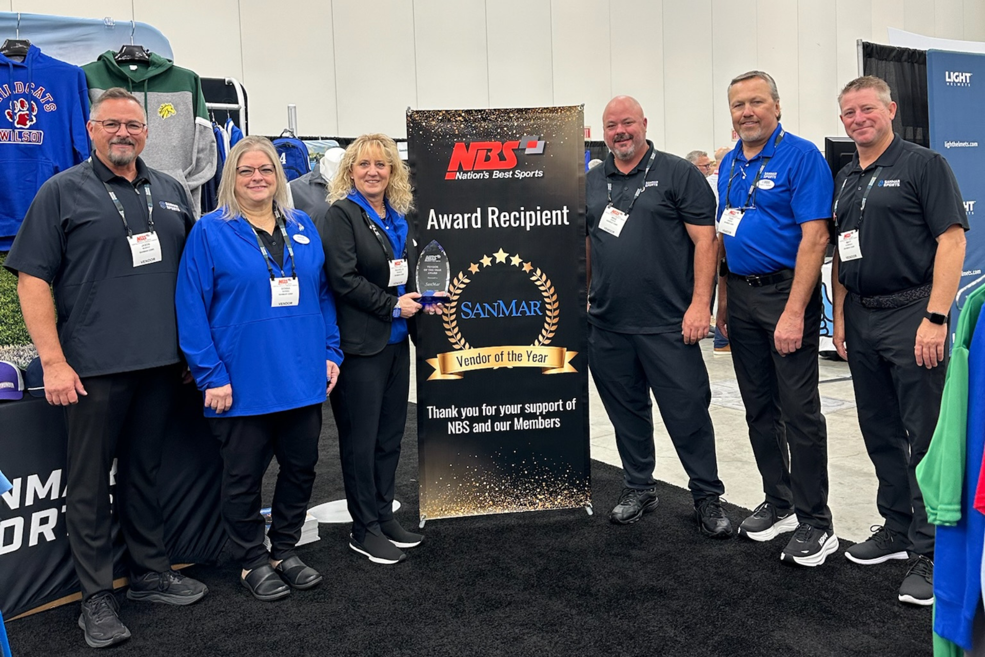 Group posing with an award trophy at an indoor event booth, smiling in blue shirts.