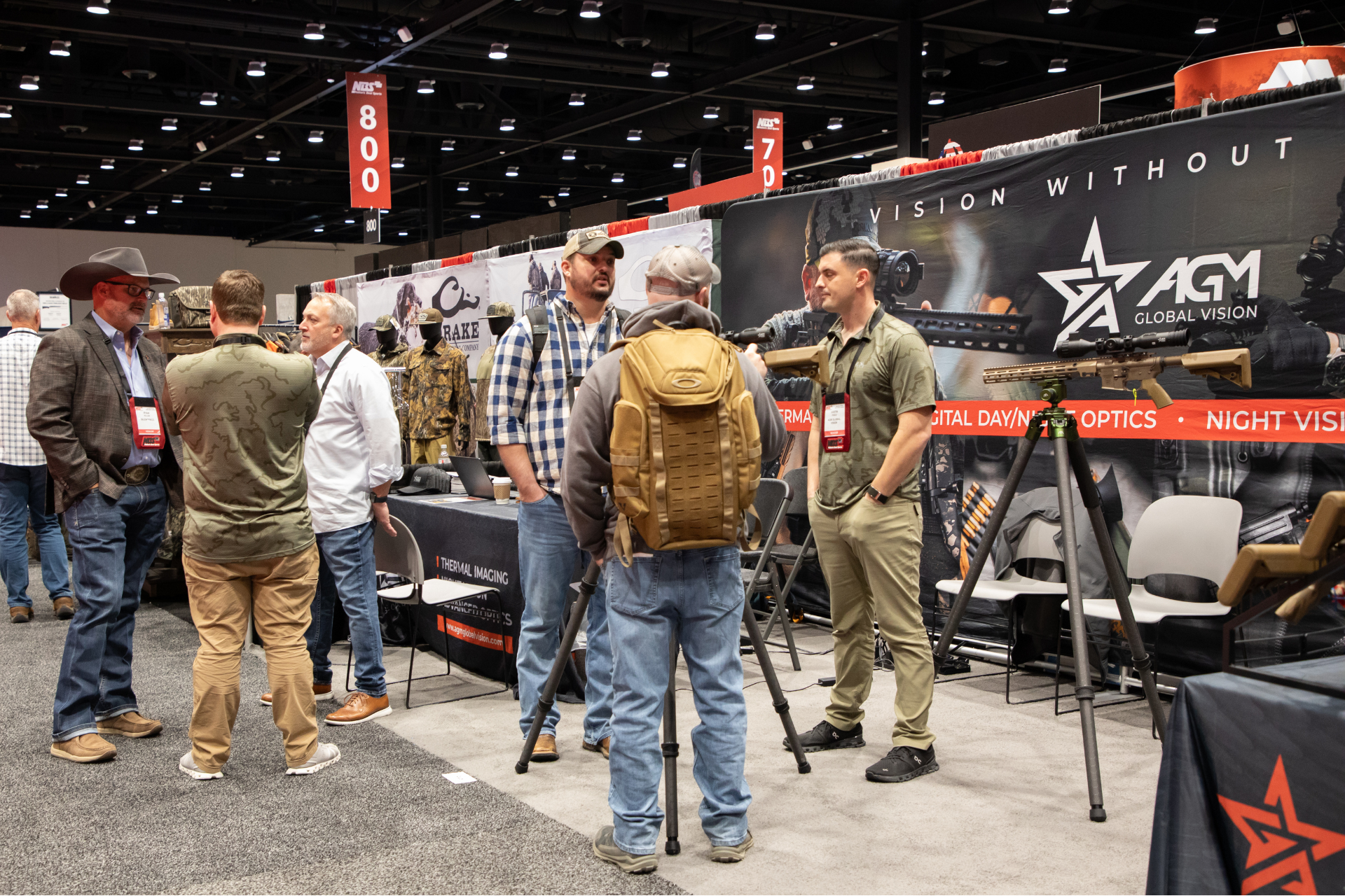 People browsing a military gear booth at a convention, with rifles and tactical bags on display.