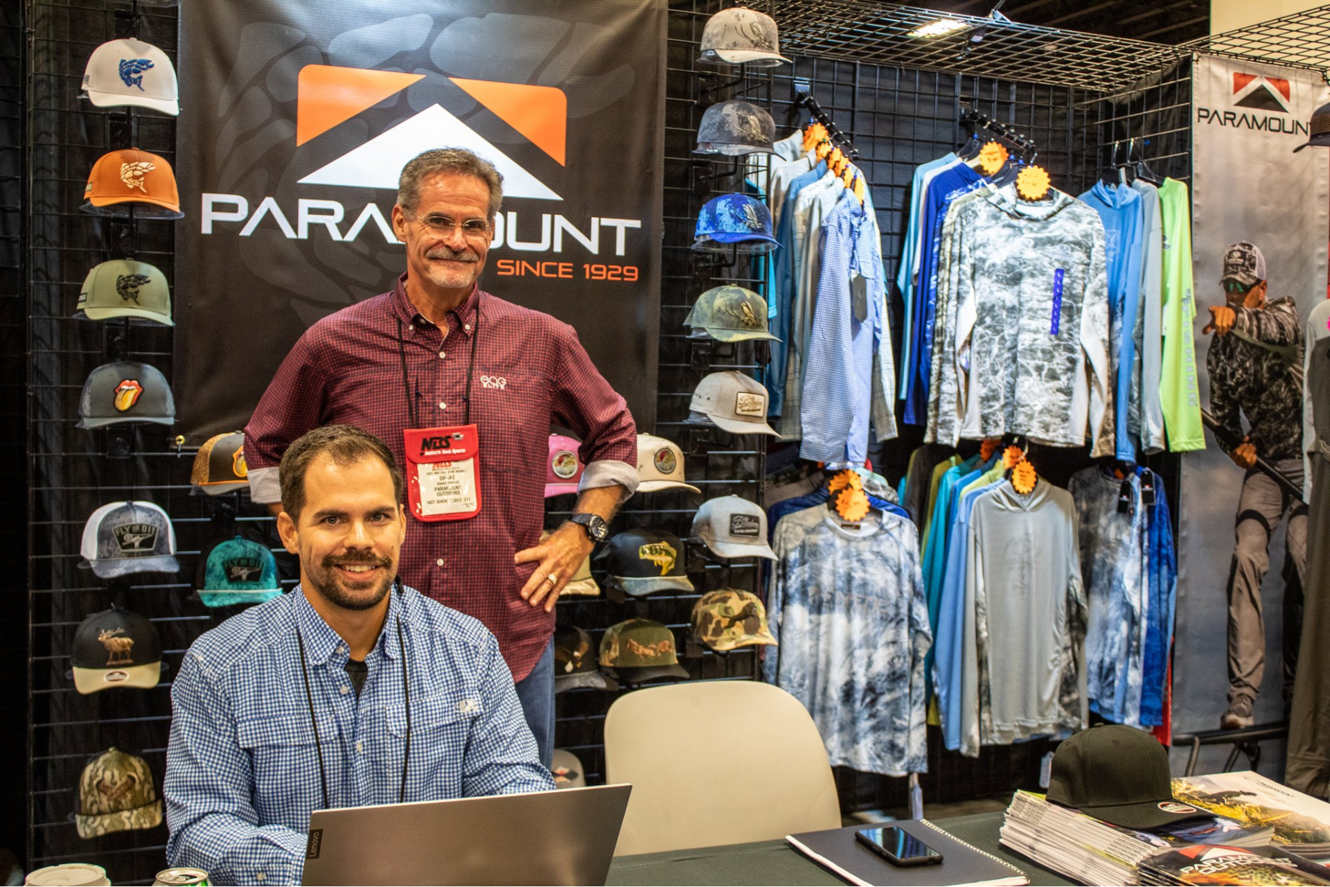 Paramount apparel booth with two men, hanging shirts, hats, and a laptop on the table