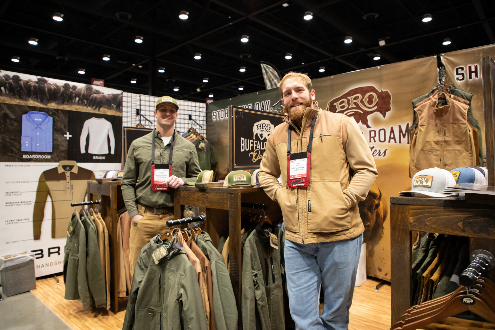 Two men posing at an outdoor clothing booth with jackets, hats, and camouflage gear on display.