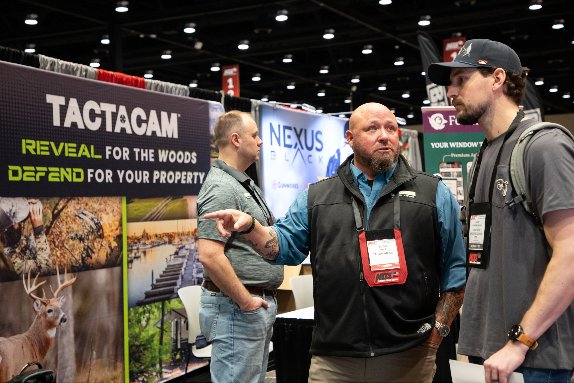 Three men talking at a trade show booth with Tactacam and Nexus signs.
