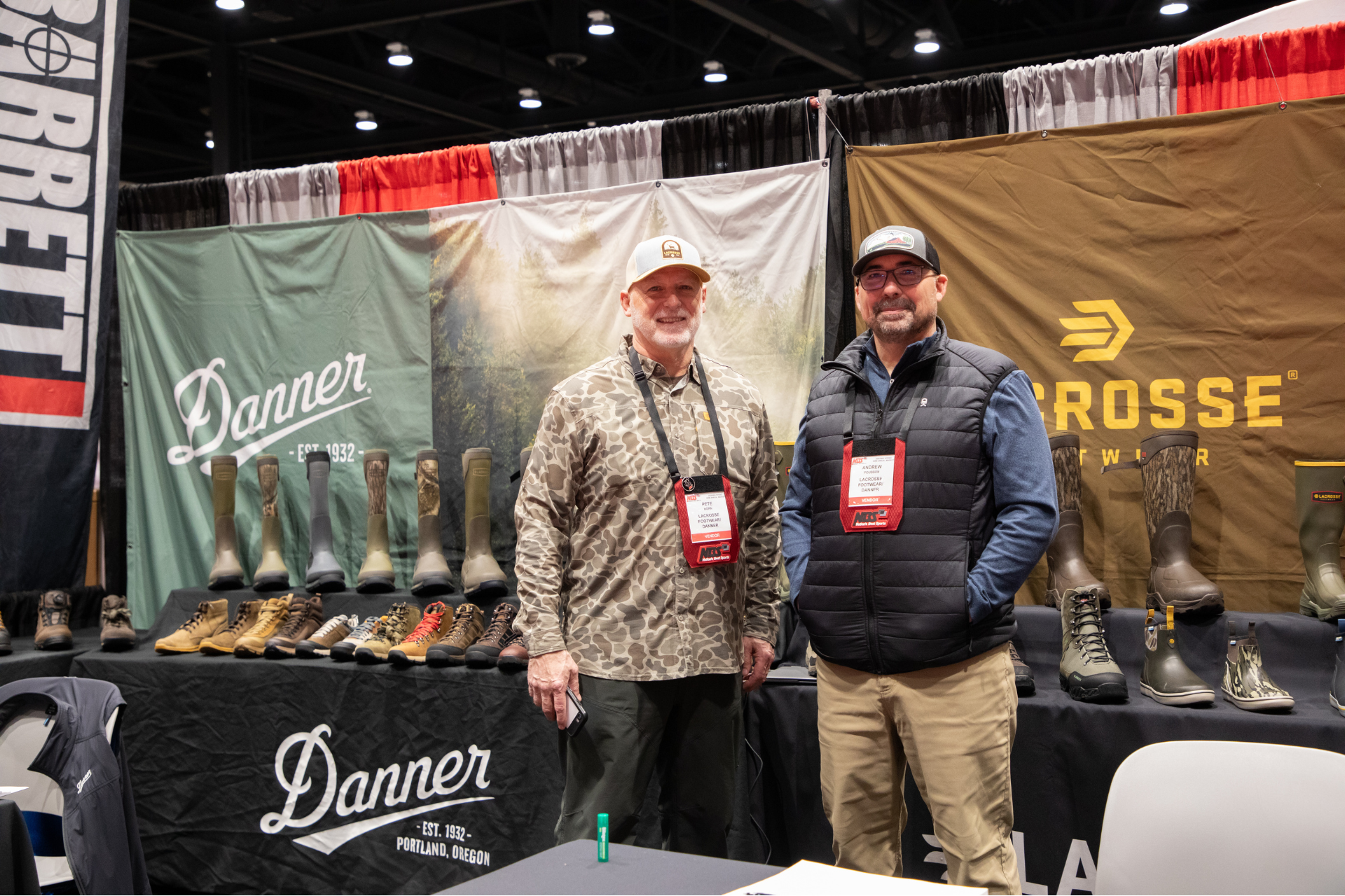 Two men stand at a Danner and Lacrosse boot booth with rows of boots displayed on a trade show table.