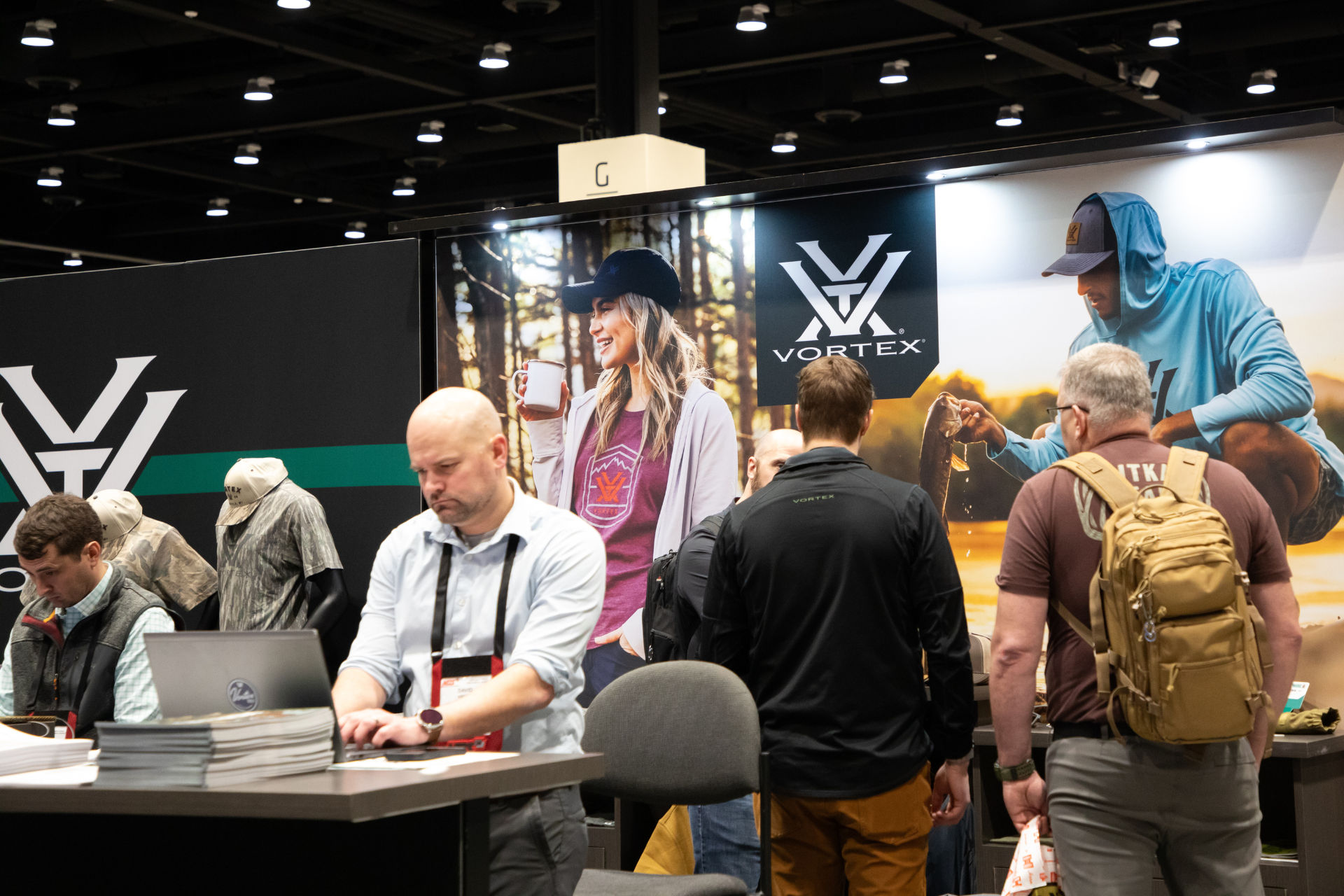 People browsing a busy indoor booth with laptops, posters, and a large V logo backdrop