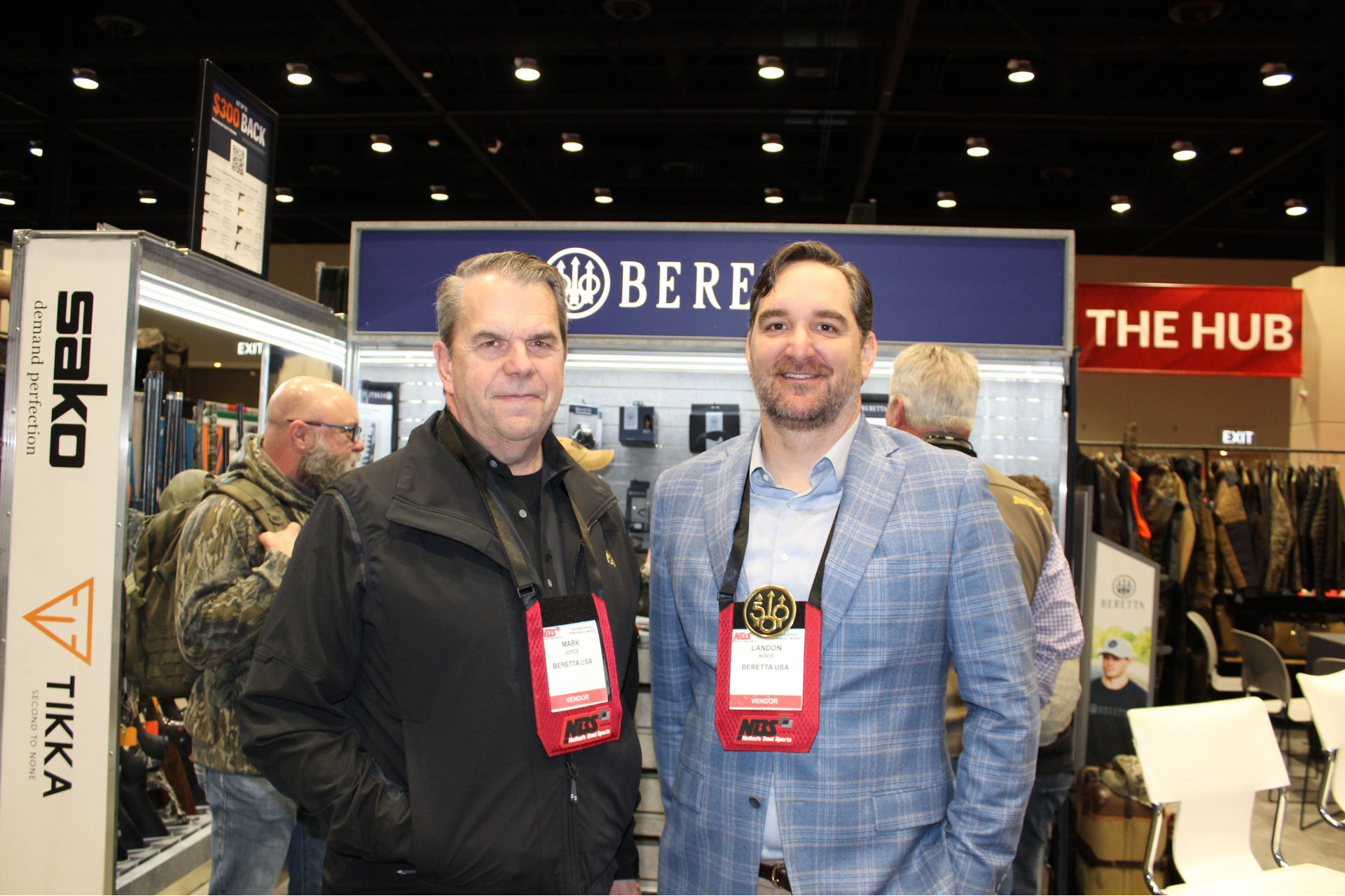 Two men posing at a trade show booth with a “BERETTA” sign and “THE HUB” banner behind them