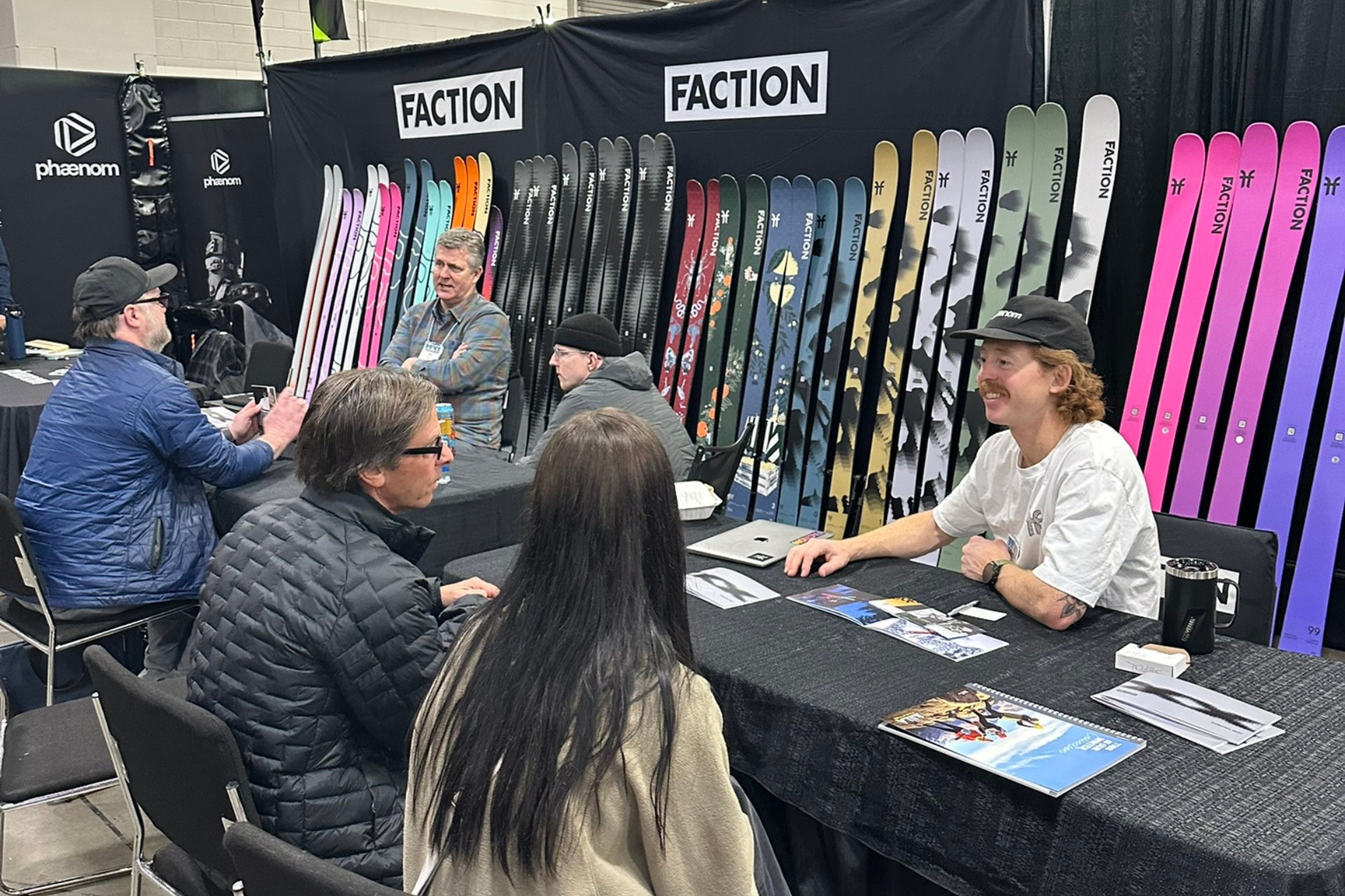 People at a ski equipment booth, with colorful skis displayed on a black Faction backdrop.