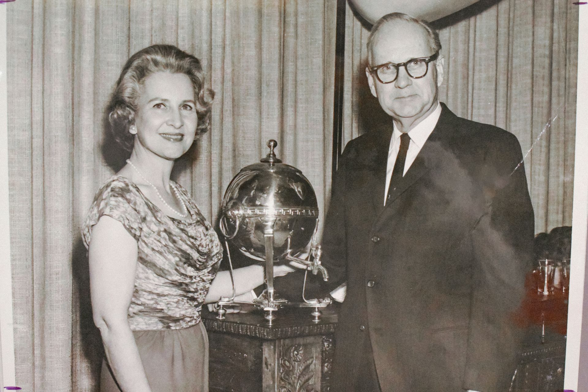 Two people standing beside a trophy-like globe on a table, with curtains in the background.