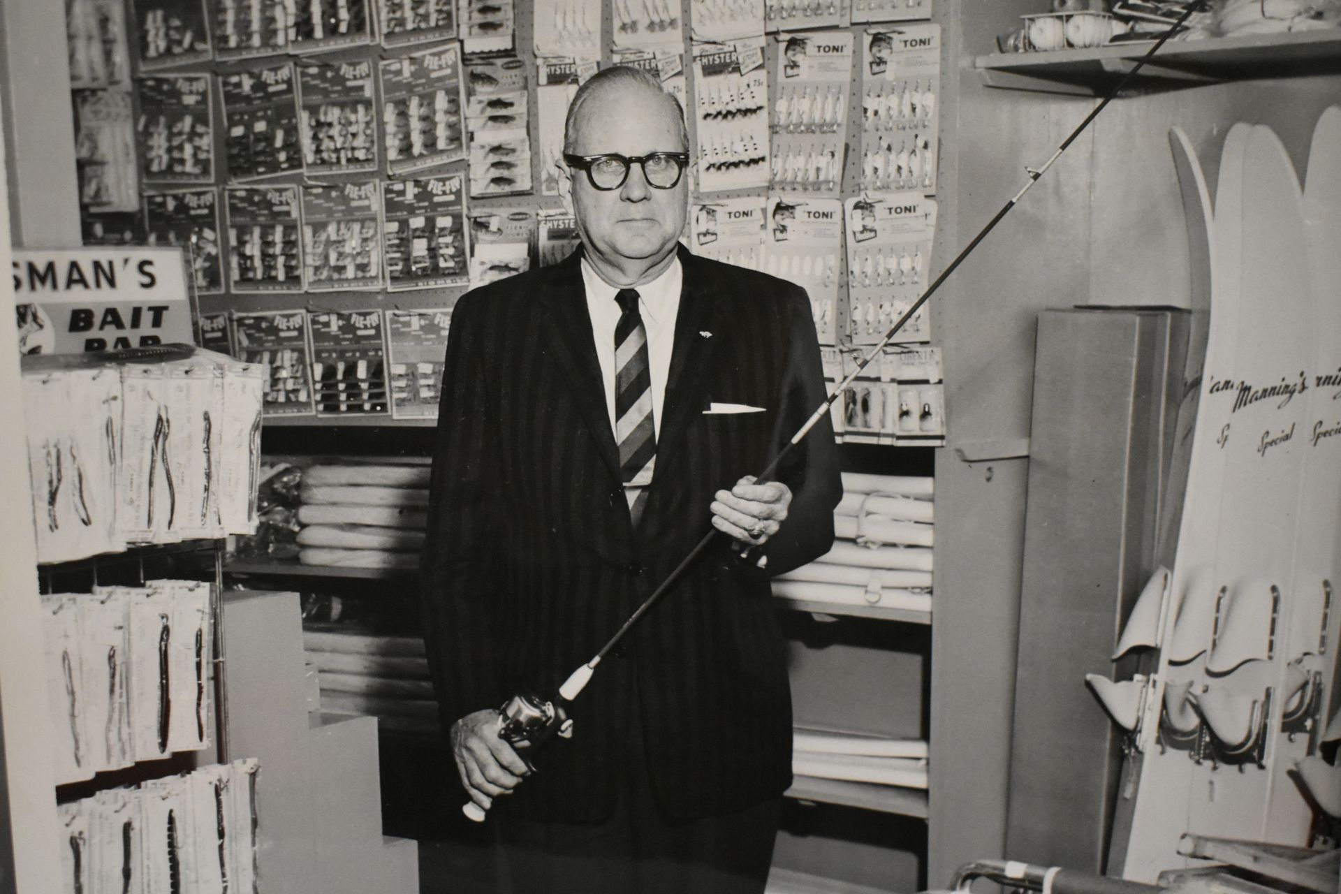 Black-and-white photo of a suited man holding a fishing rod in a tackle shop