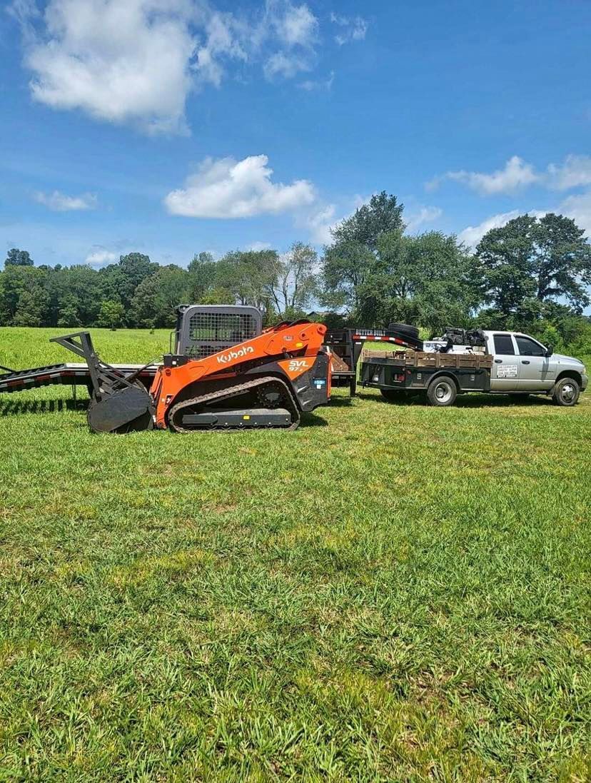 An orange skid steer loader sits on a grassy field next to a silver flatbed truck under a blue sky.