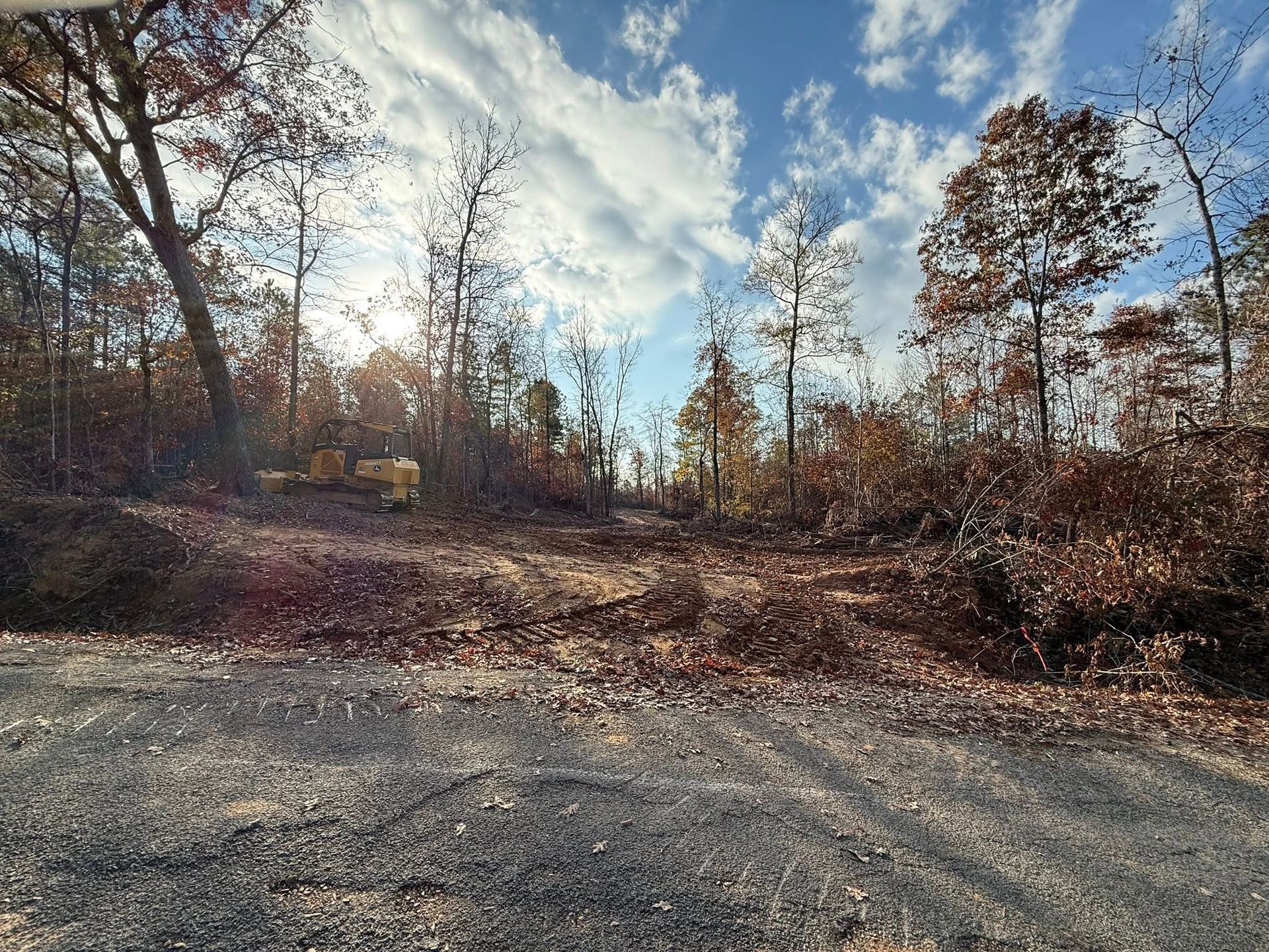 A yellow piece of construction equipment clears a path through a forest under a bright, partly cloudy blue sky.