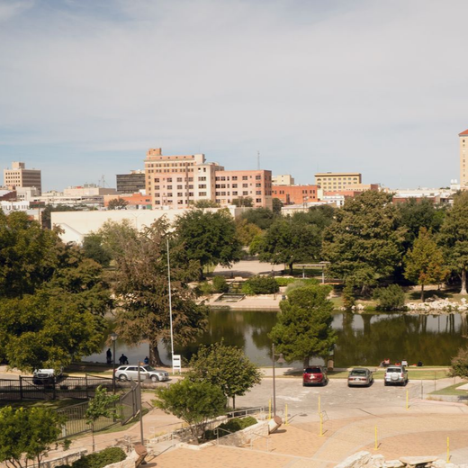 City skyline beyond a tree-lined park with a pond and parked cars in the foreground