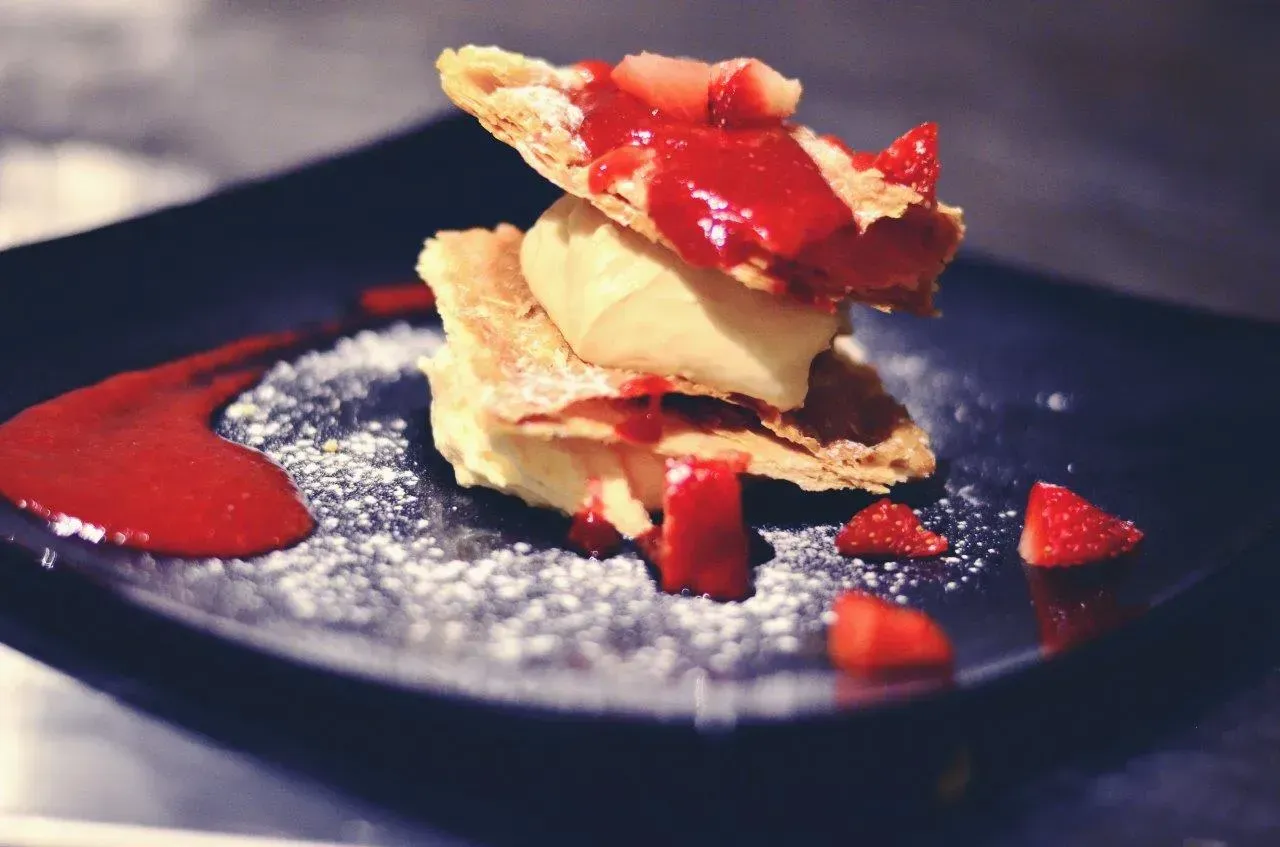 Dessert millefoglie di fragole disposto su un piatto nero, condito con salsa di fragole e spolverato con zucchero a velo.