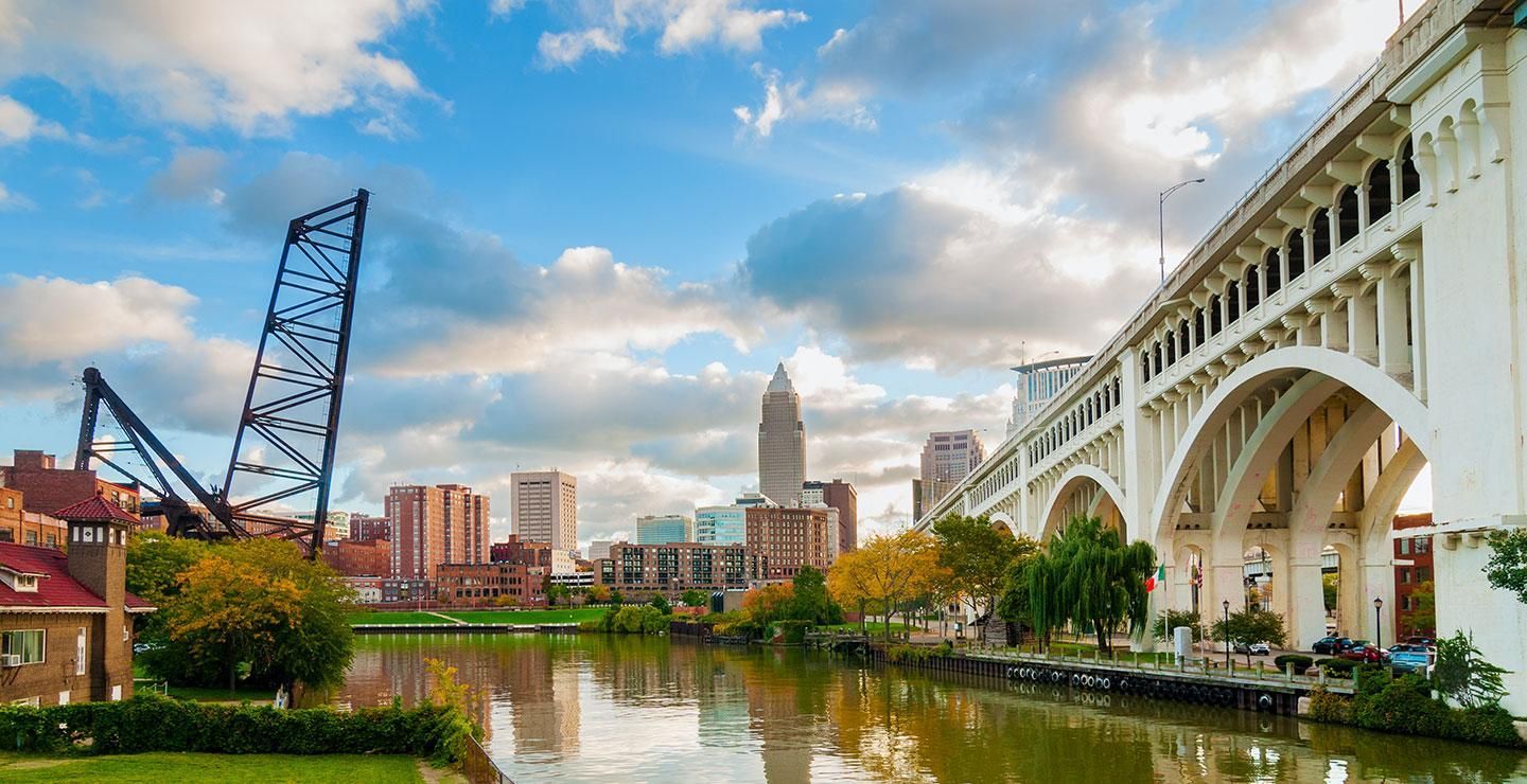 City skyline with a river, white arched bridge, and cloudy sky at sunset