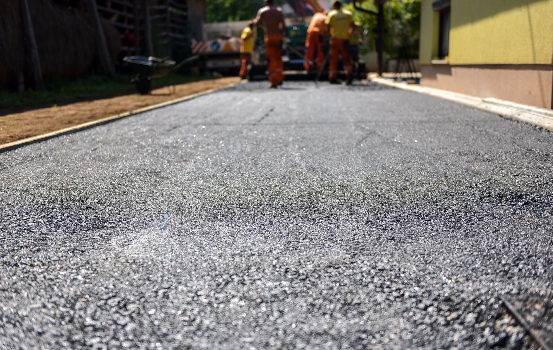 Workers paving a road with asphalt; blurred figures in orange vests in the background.