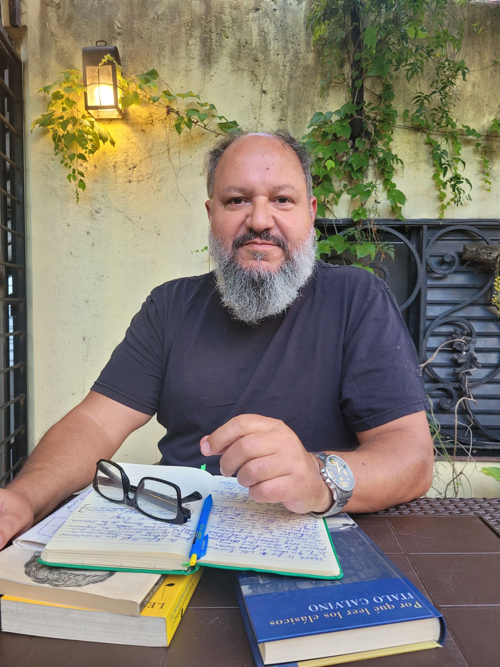 Un hombre sentado en una mesa al aire libre, sonriendo, con libros, un cuaderno y gafas delante de él.
