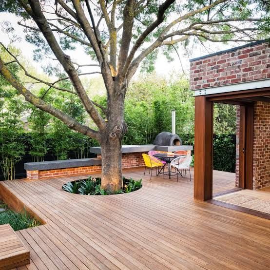 Modern wooden deck with a large tree, outdoor seating, and brick patio beside a house