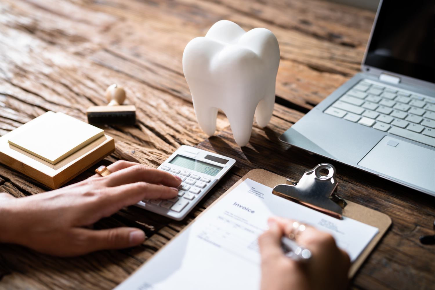 Hands using a calculator beside a clipboard, laptop, and tooth model on a wooden desk