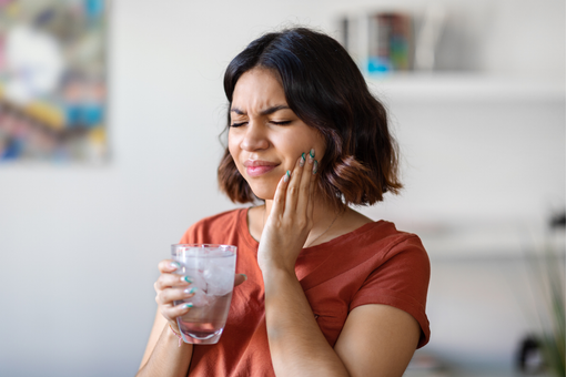 Woman holding a glass of water and touching her cheek, looking pained in a bright room