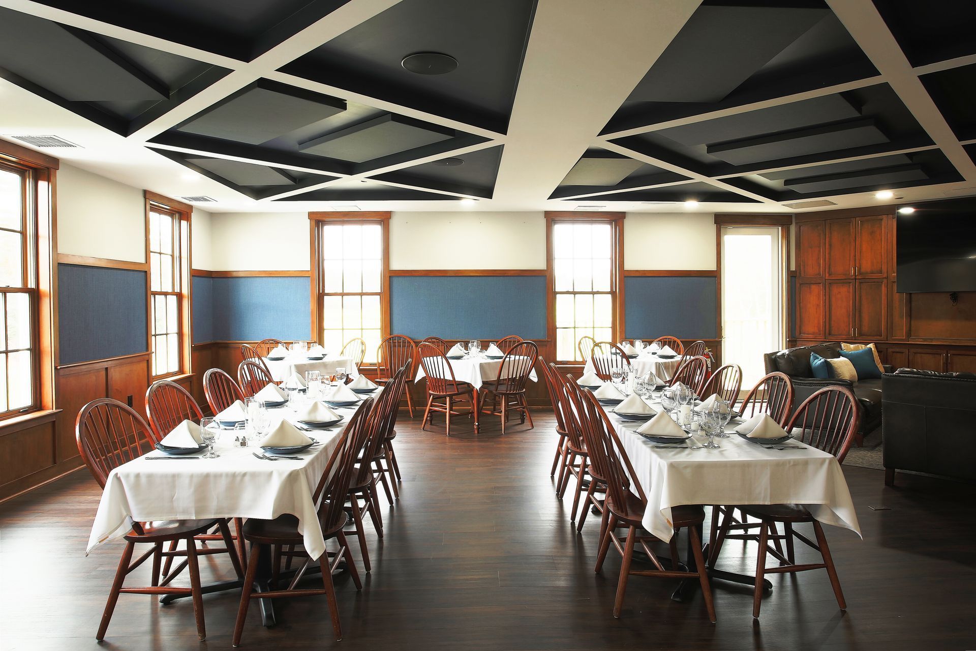 Dining room with long tables set for a meal, wooden floors and walls, blue accents, and a geometric ceiling.