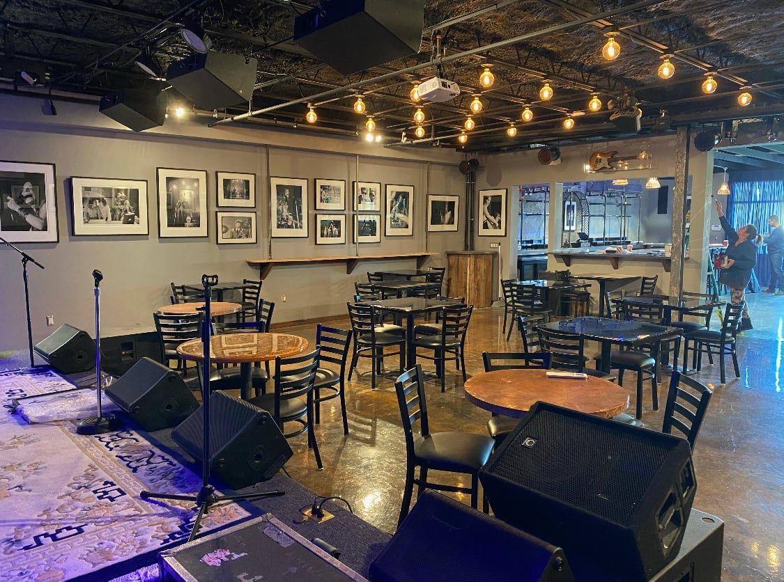 Interior of a music venue with tables, stage, sound equipment, and art on the wall.