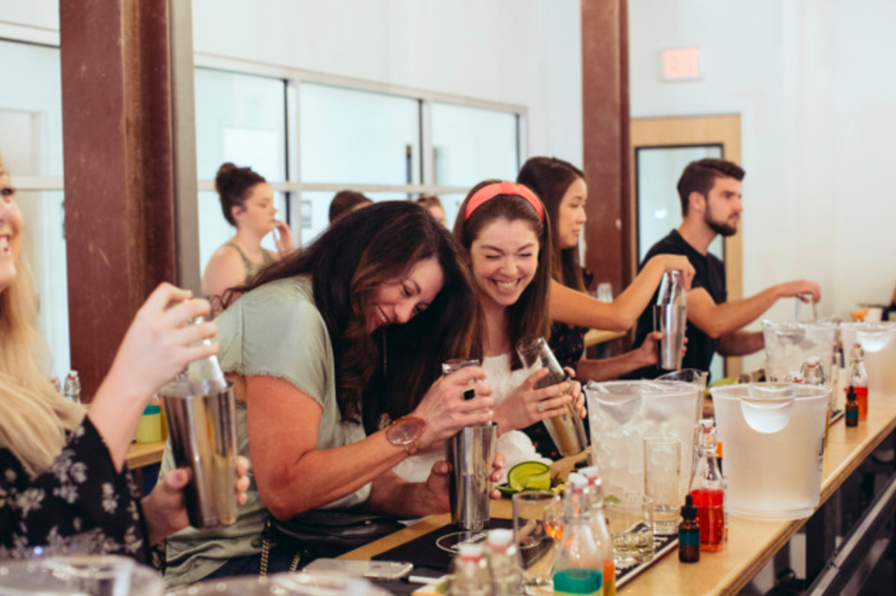 People laughing while mixing cocktails at a bar. Some wearing aprons, with drinks and ice.