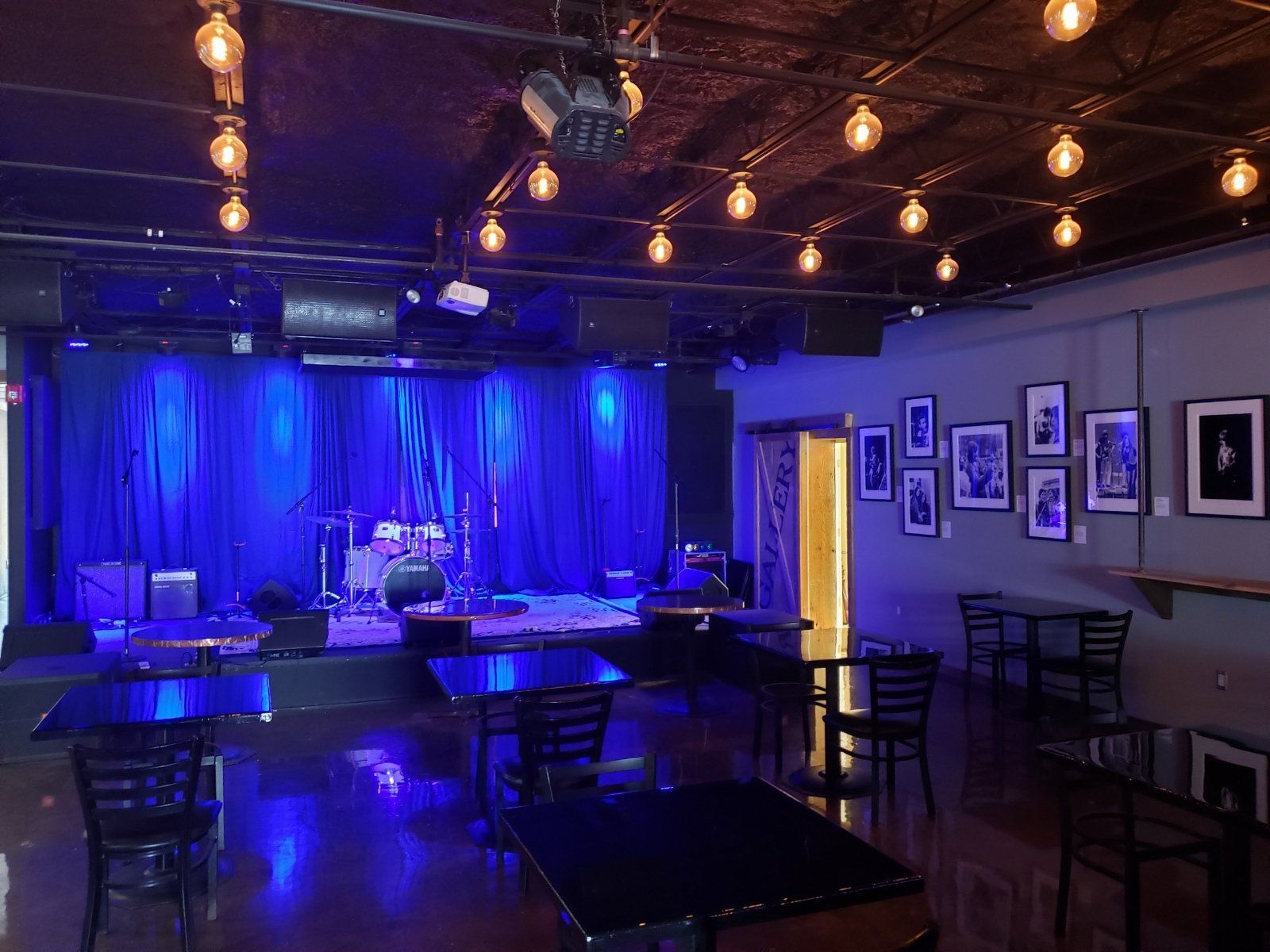 Empty music venue with a stage, tables, chairs, and art on the wall, illuminated with blue lights.