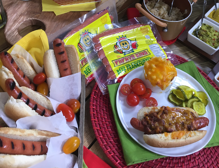Hot dogs, buns, tomatoes, pickles, and mac and cheese on a table setting, with packages of hot dogs.