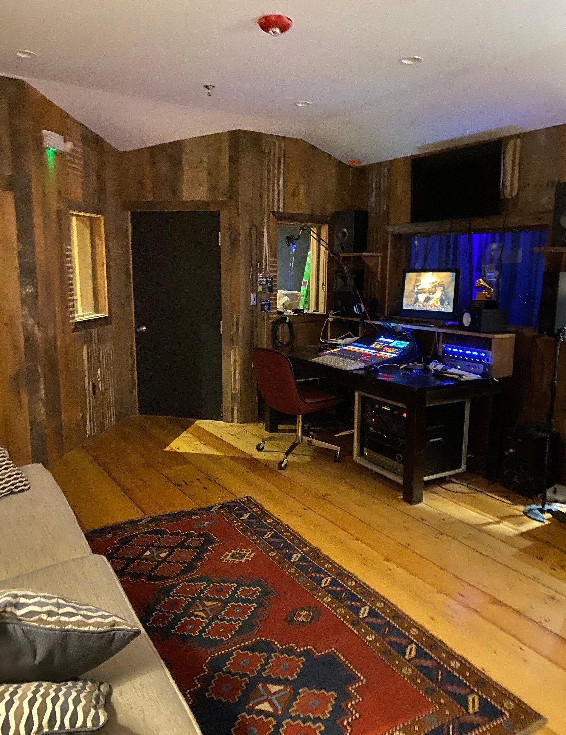 Recording studio interior: wood-paneled walls, desk with equipment, rug, couch, and a dark door.