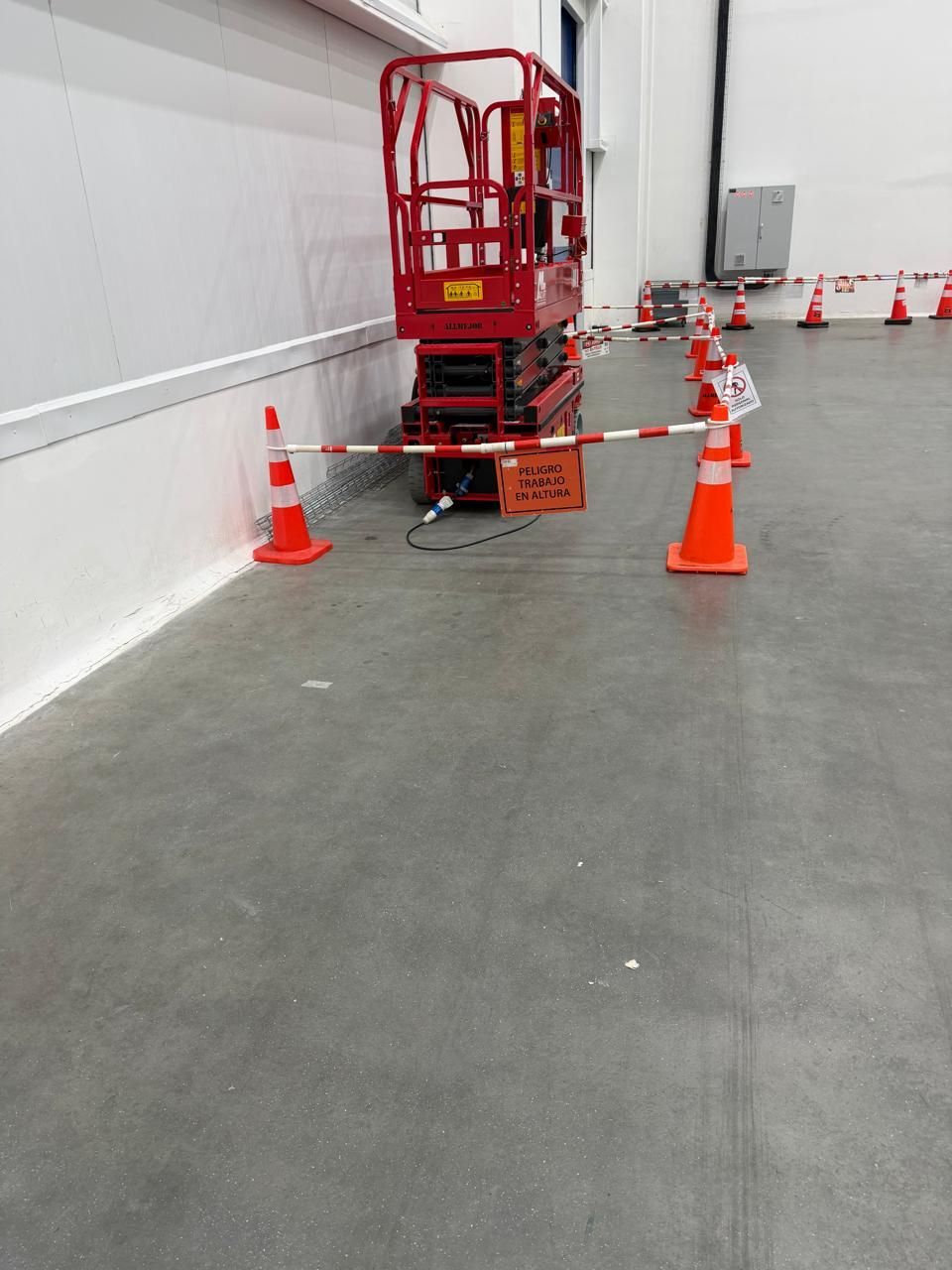 Una plataforma elevadora de tijera roja rodeada de conos naranjas en el pasillo de un almacén.