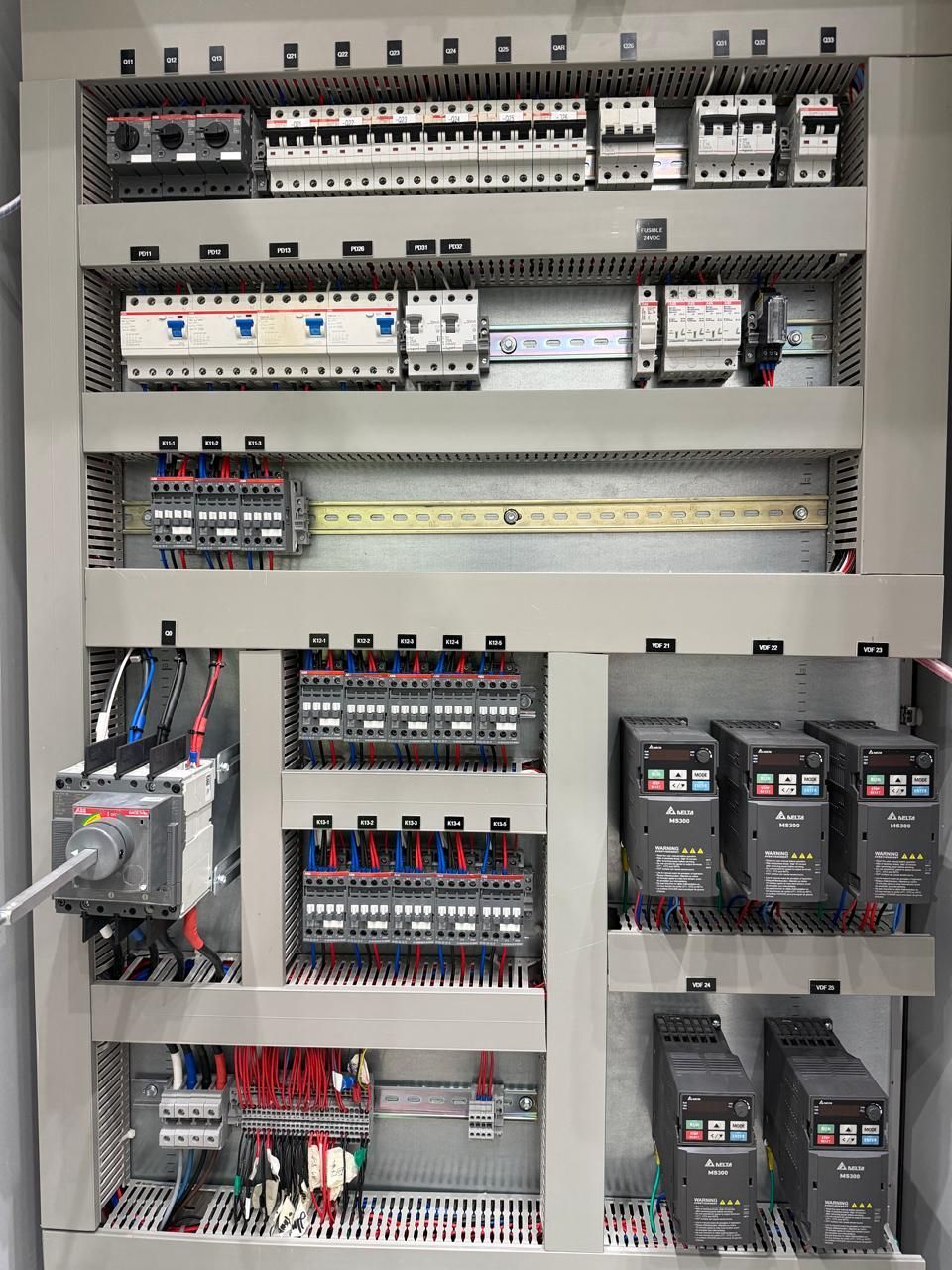 Panel de control eléctrico abierto con filas de interruptores, relés, cableado y variadores dentro de un gabinete metálico.