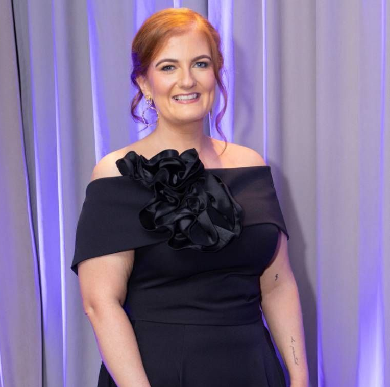 Woman in a black off-shoulder dress with ruffled detail, standing before purple drapes and smiling.