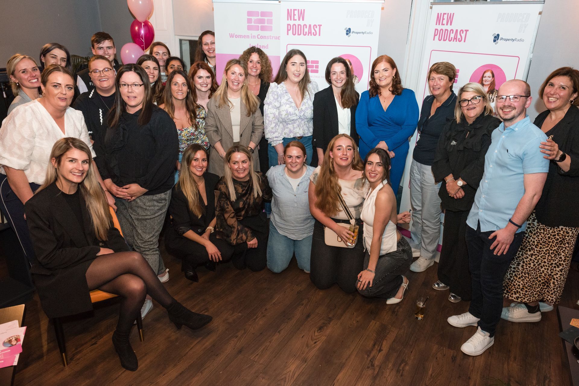 Group posing indoors at a pink-themed event backdrop, smiling and standing closely together.