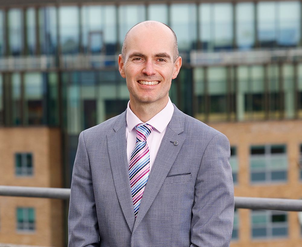 Man in a gray suit and striped tie standing outdoors in front of a modern office building.