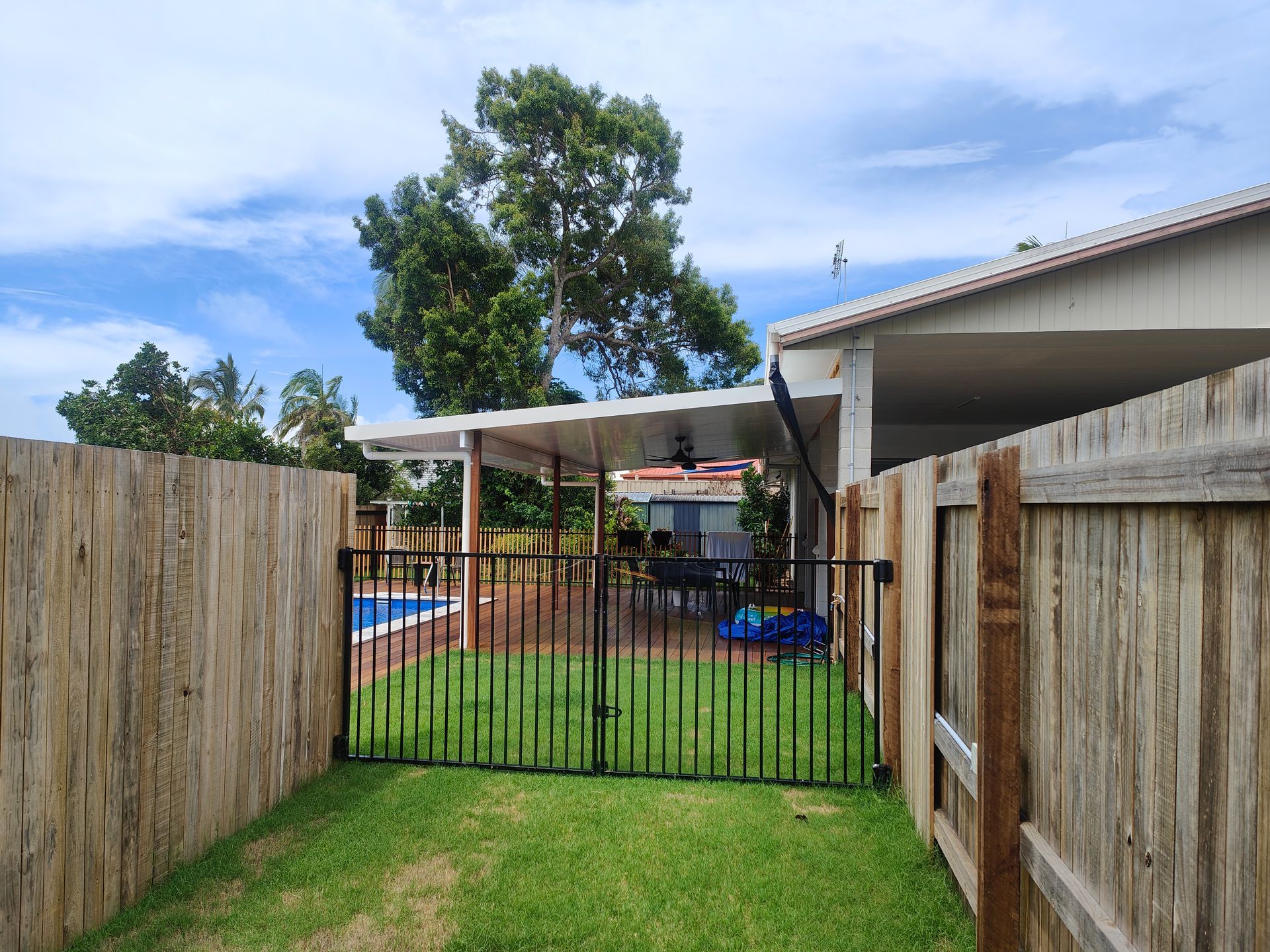 Backyard With A Pool, Grassy Lawn, Wooden Fences, And A Black Gate Under A Partly Cloudy Sky — Quality Fences 4 U in Gympie, QLD