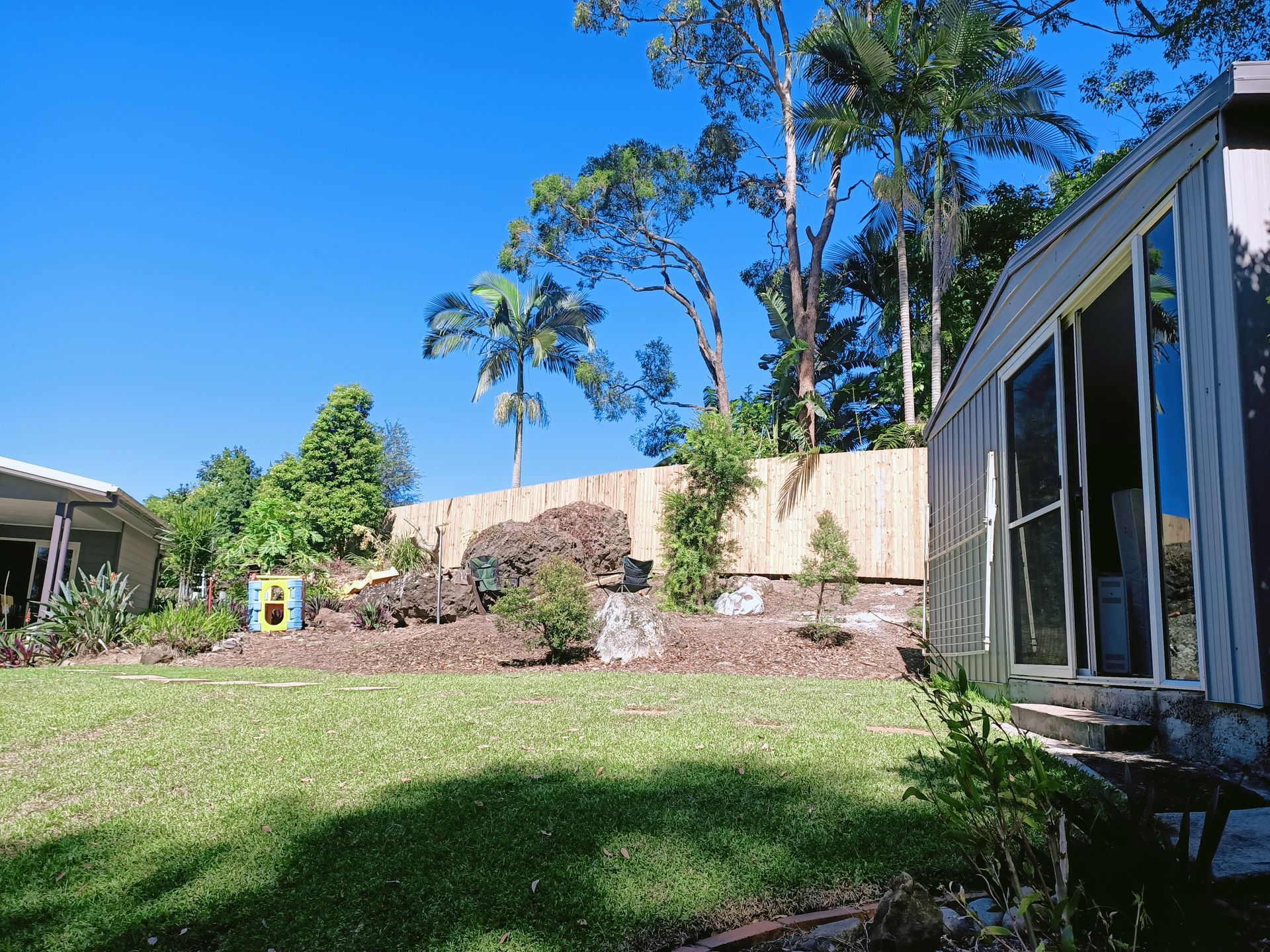 Sunny Backyard With Green Lawn, Stone Retaining Wall, Tall Trees, And A Modern House Exterior — Quality Fences 4 U in Gympie, QLD